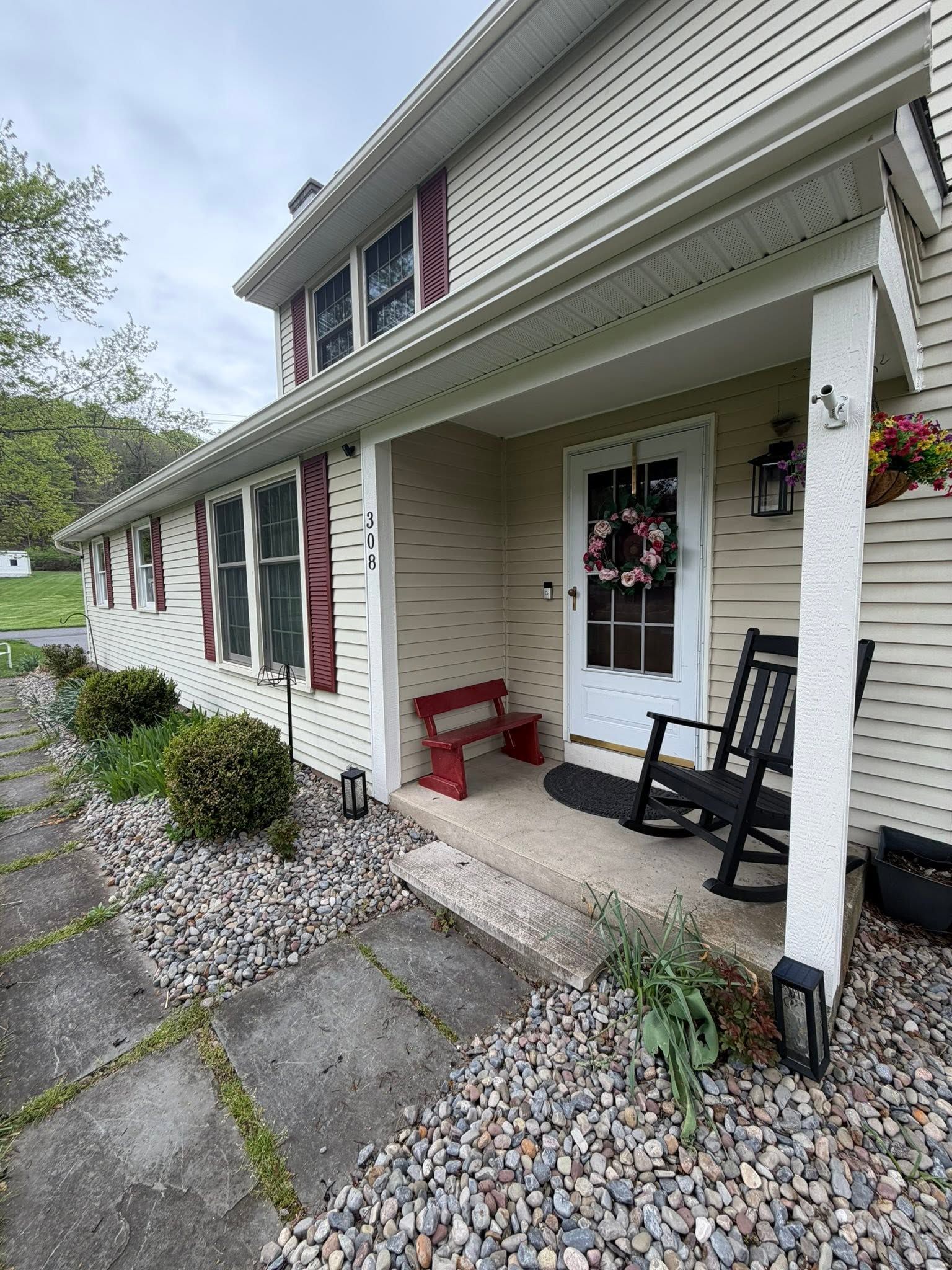 Front porch of a beige house with red shutters, white door, black rocking chair, and gravel walkway.