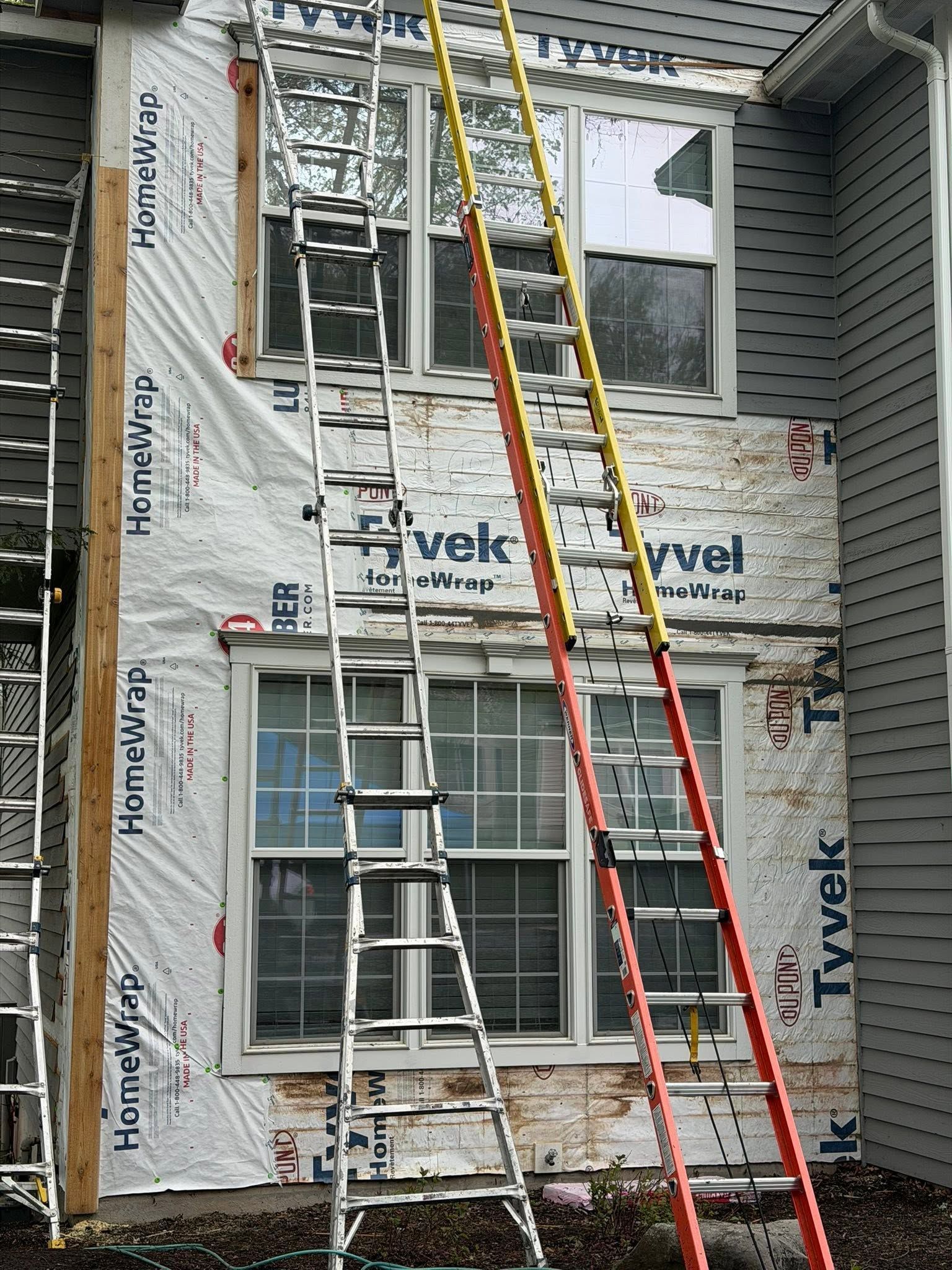Two ladders lean against a house under renovation with Tyvek wrap and exposed windows.