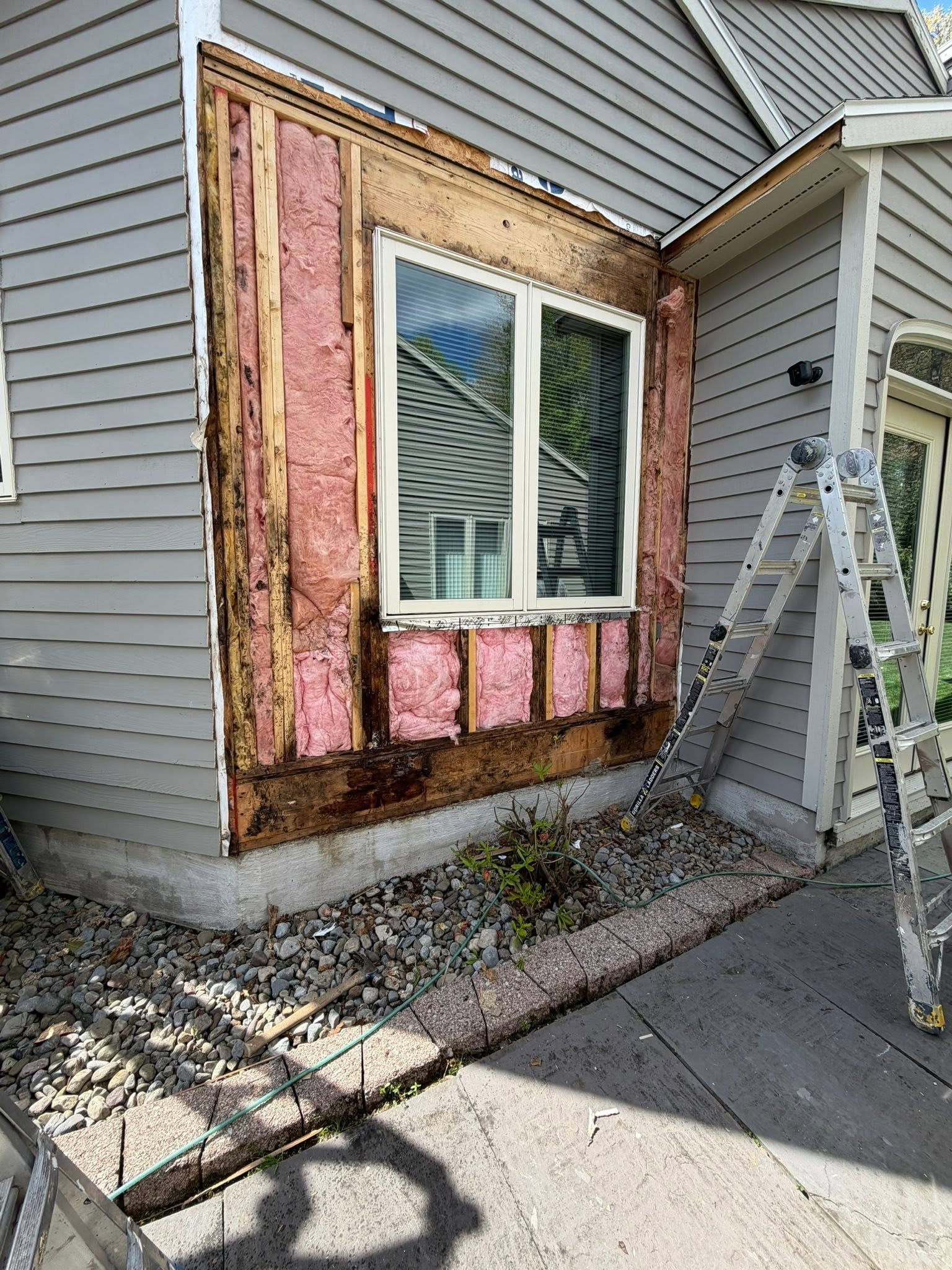 Damaged house wall with exposed pink insulation around a window, next to a ladder and siding repair area.