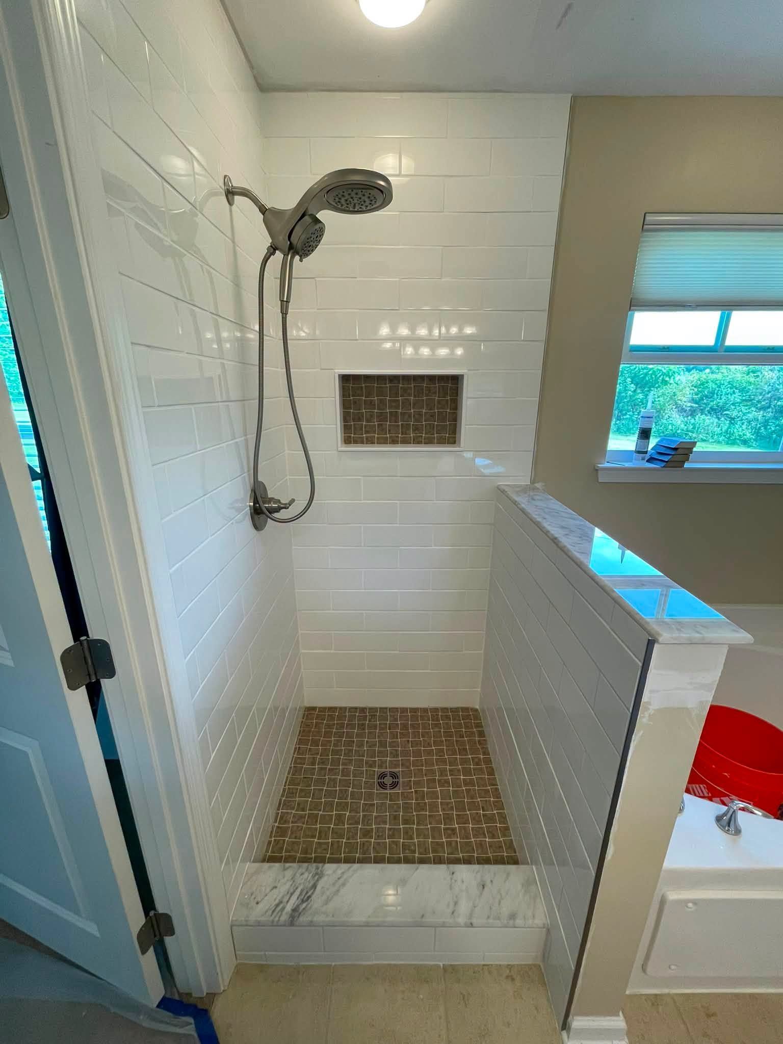 Small white-tiled shower with glass door, handheld showerhead, built-in niche, and pebble floor.