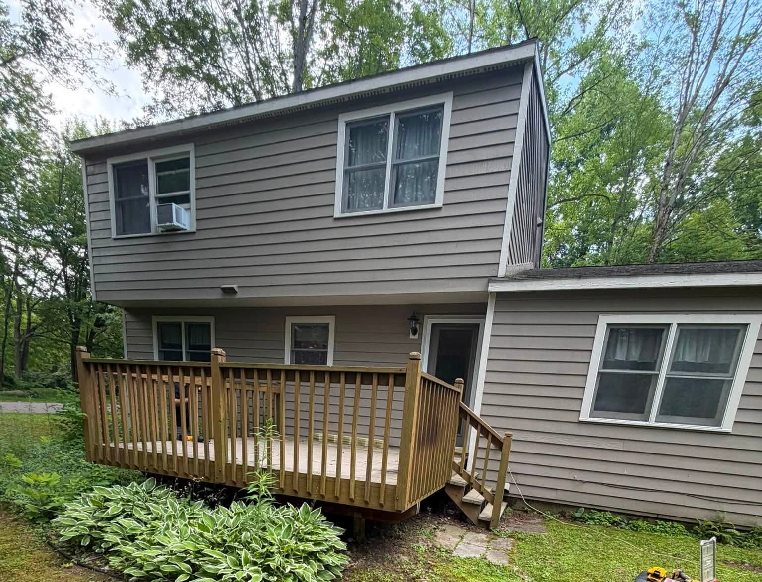Two-story beige house with a wooden deck and stairs, surrounded by trees and greenery.