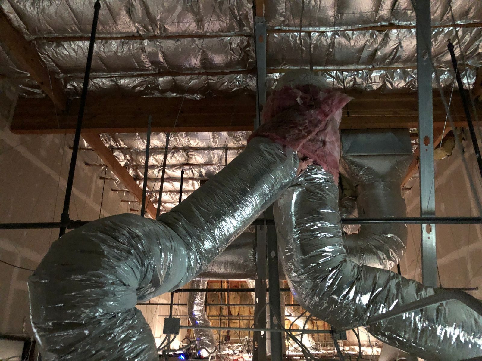 Flexible silver HVAC ducts hanging from a ceiling in an unfinished attic with exposed insulation.