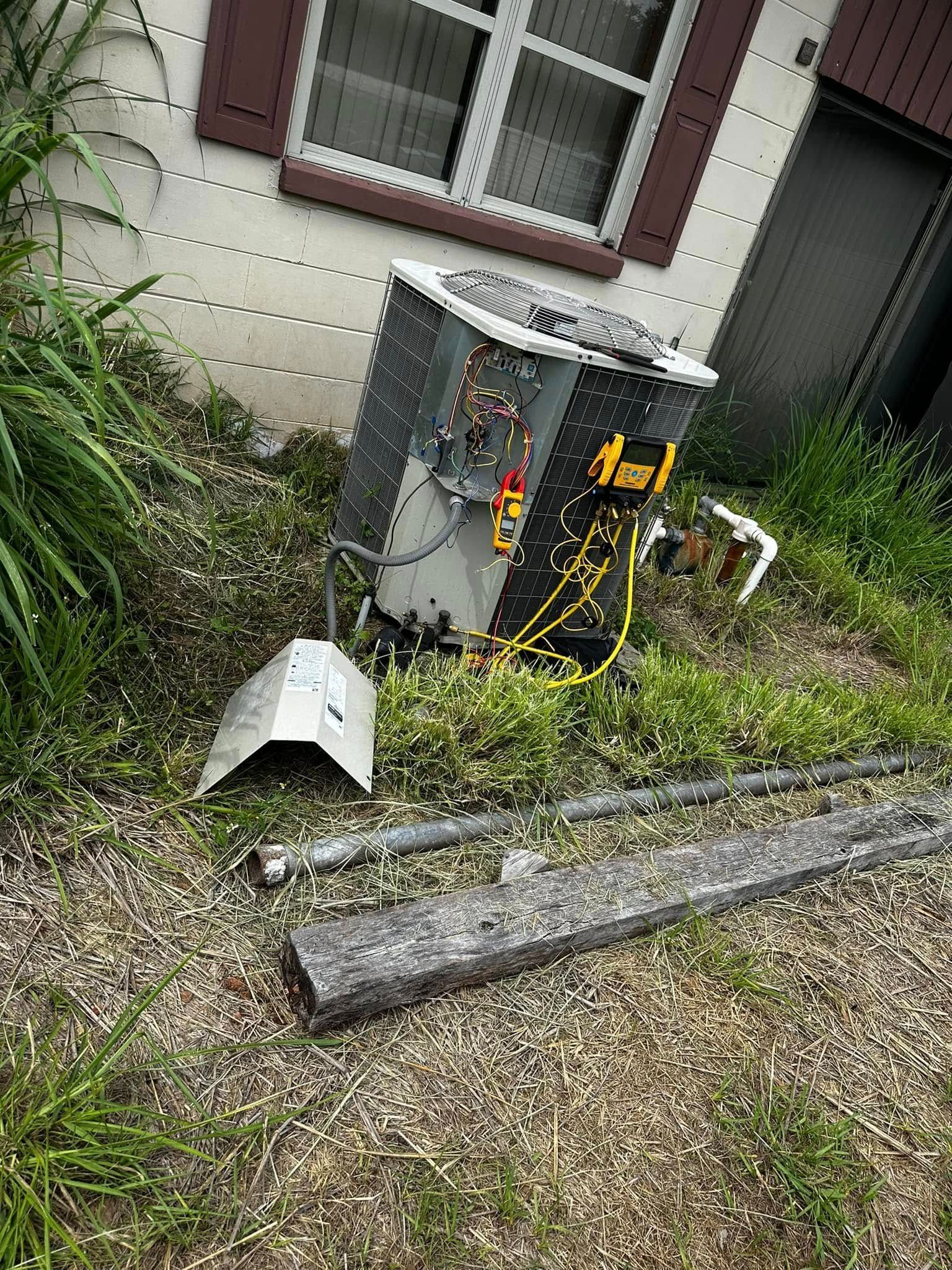 Outdoor air conditioner unit beside a house, with weeds and a detached vent cover on the ground