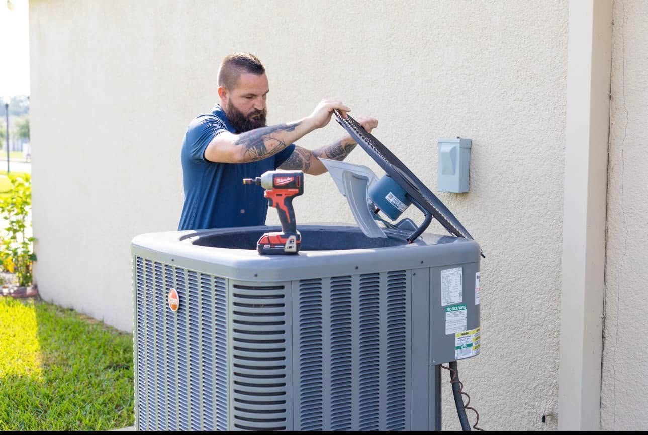 Man repairing an outdoor air conditioner unit with a power tool beside a house.