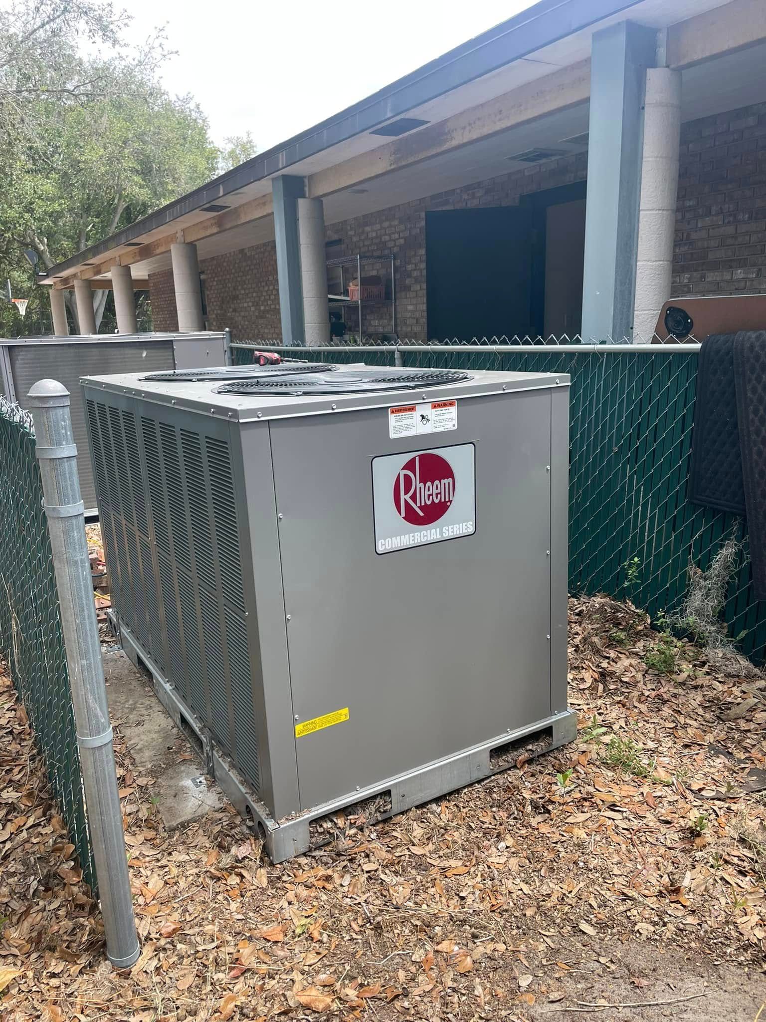 Gray HVAC unit beside a house, next to a green chain-link fence and under a covered porch.
