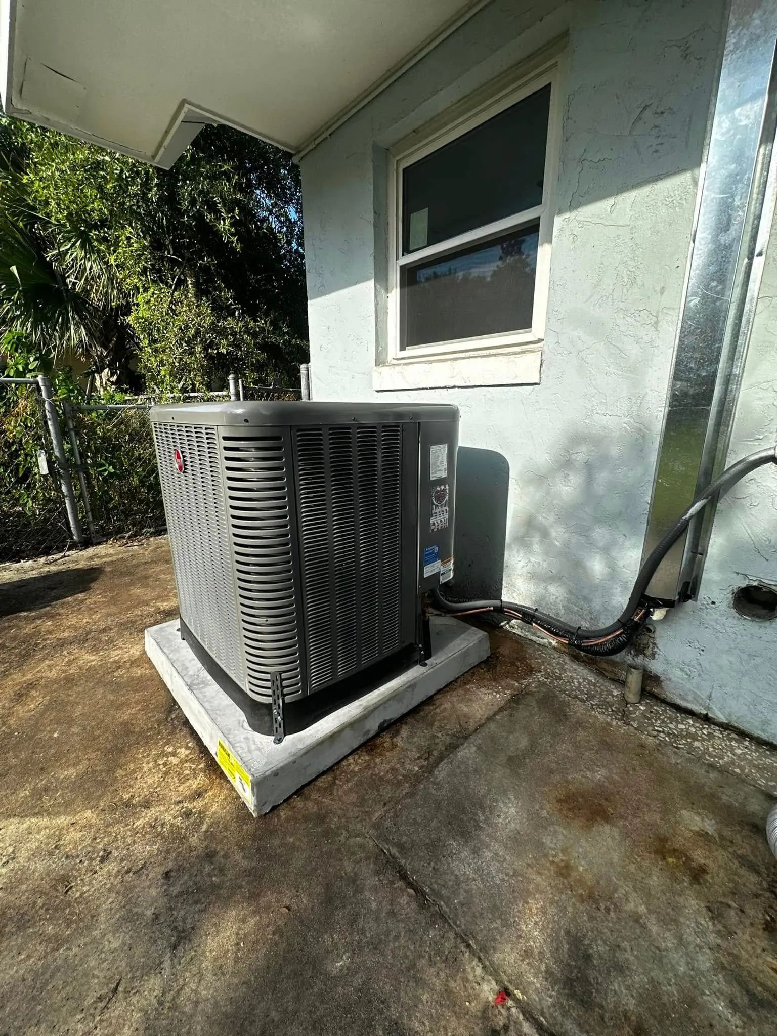 Outdoor air conditioning unit beside a house window, with black fan grille and sunlit concrete pad
