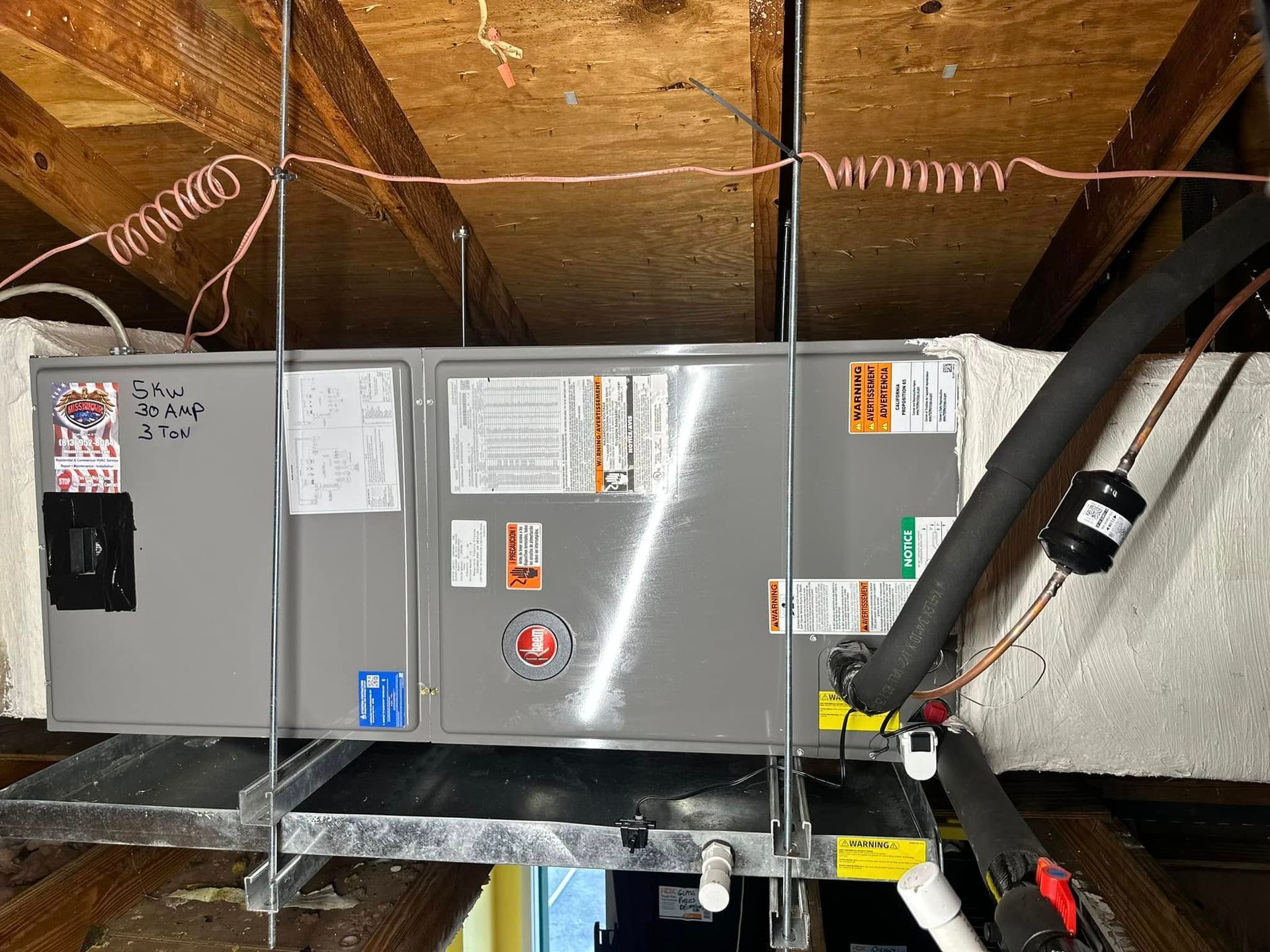 Gray HVAC unit in an attic, with ductwork and wiring under the wooden roof rafters.