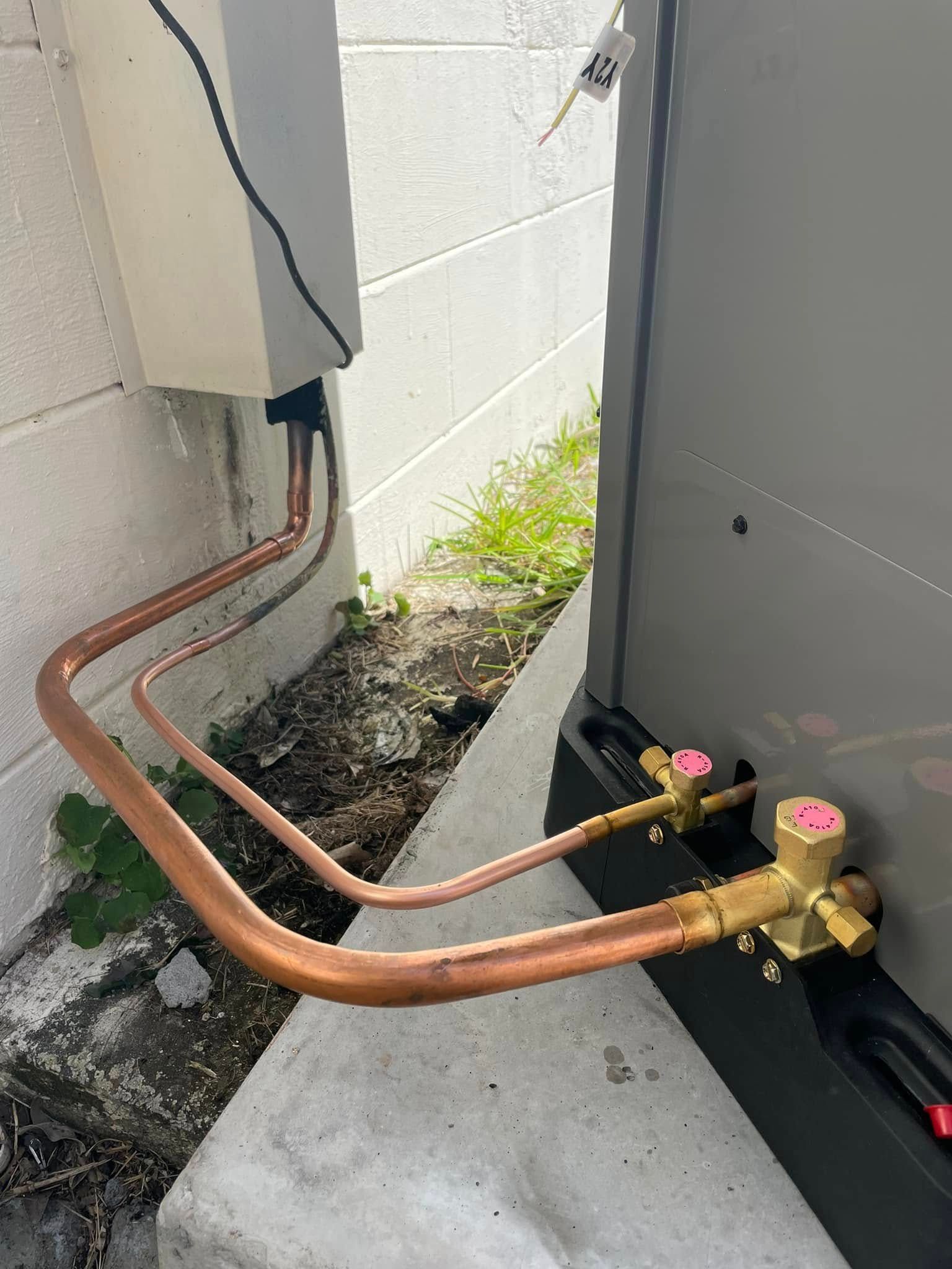 Copper pipes connected to a gray outdoor unit beside a white wall on a concrete slab