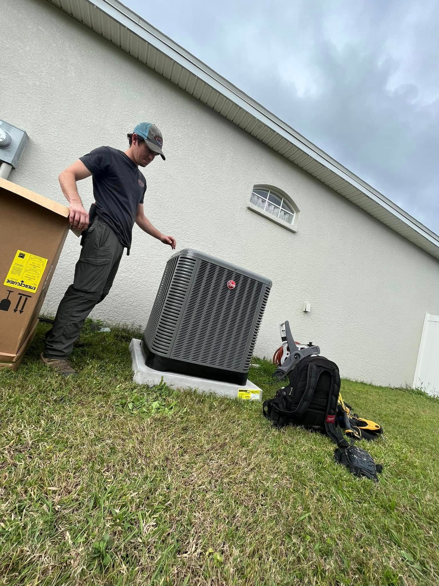 Man standing beside an outdoor HVAC unit and tools near a house wall under cloudy sky