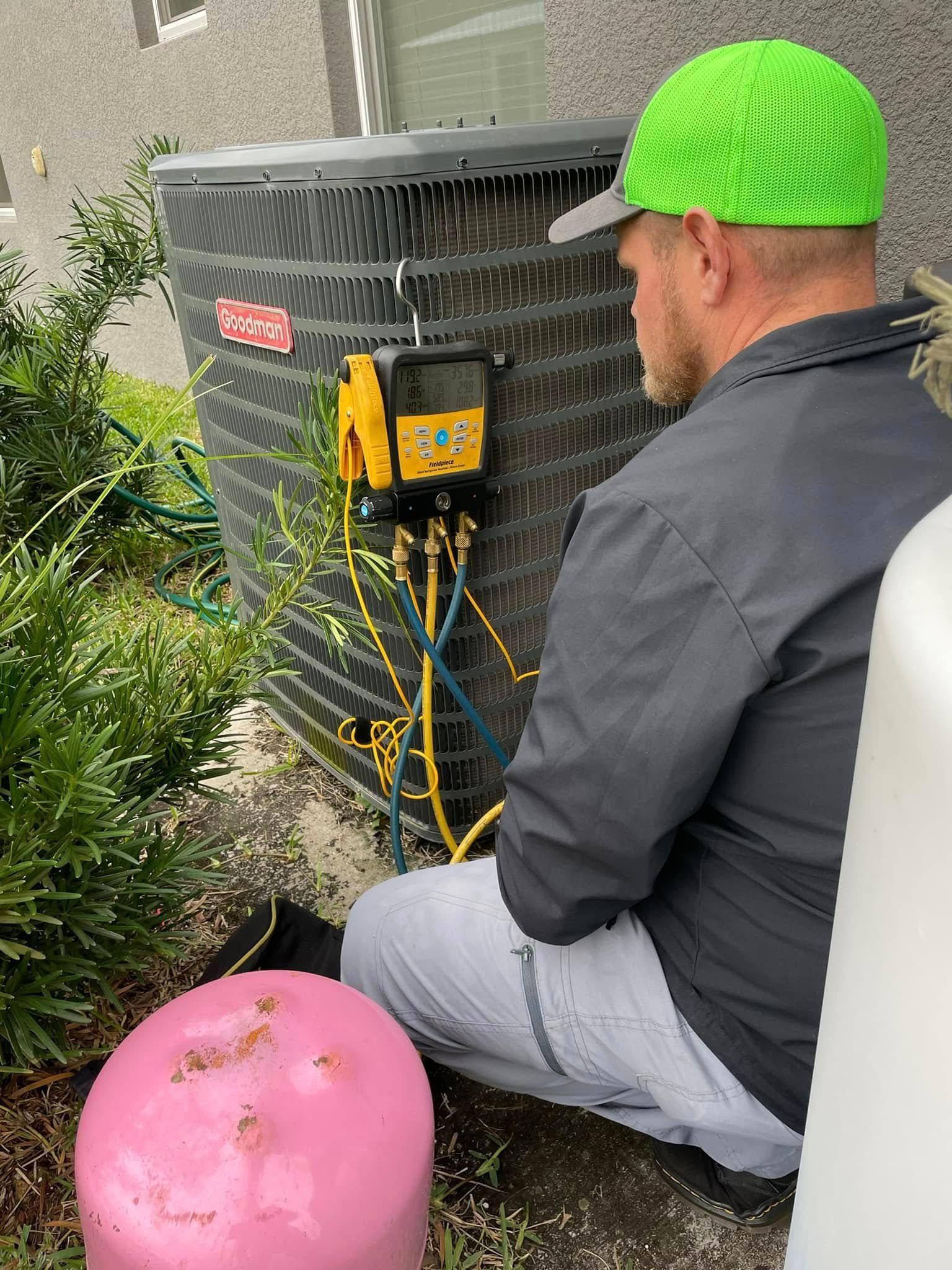 Man kneeling beside outdoor HVAC unit, wearing a neon green cap and black jacket, with yellow wires connected.