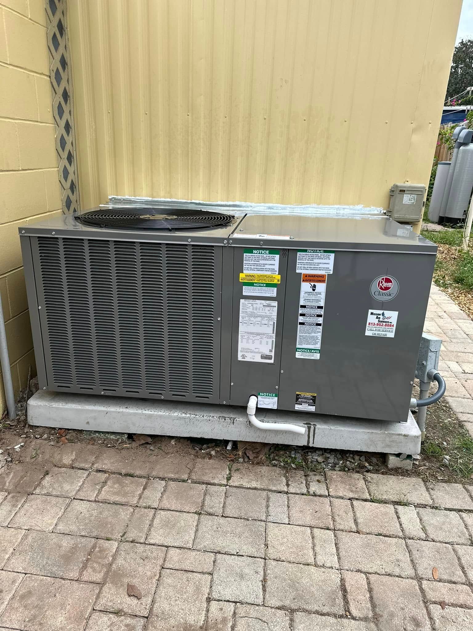 Outdoor HVAC unit beside a yellow wall, with dark gray vented front and warning labels.