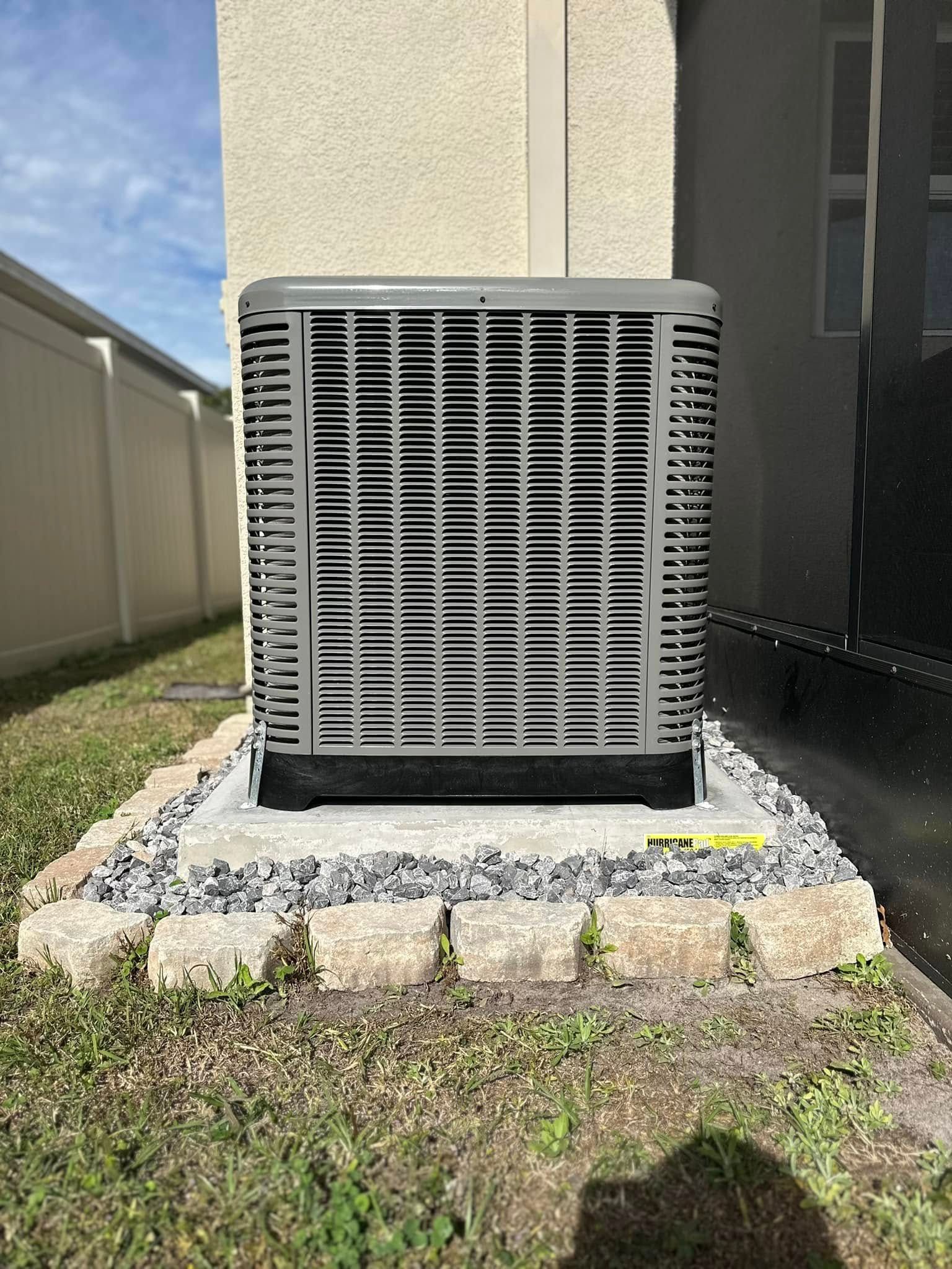 Outdoor HVAC unit beside a house, on a small gravel pad near a wall and fence.