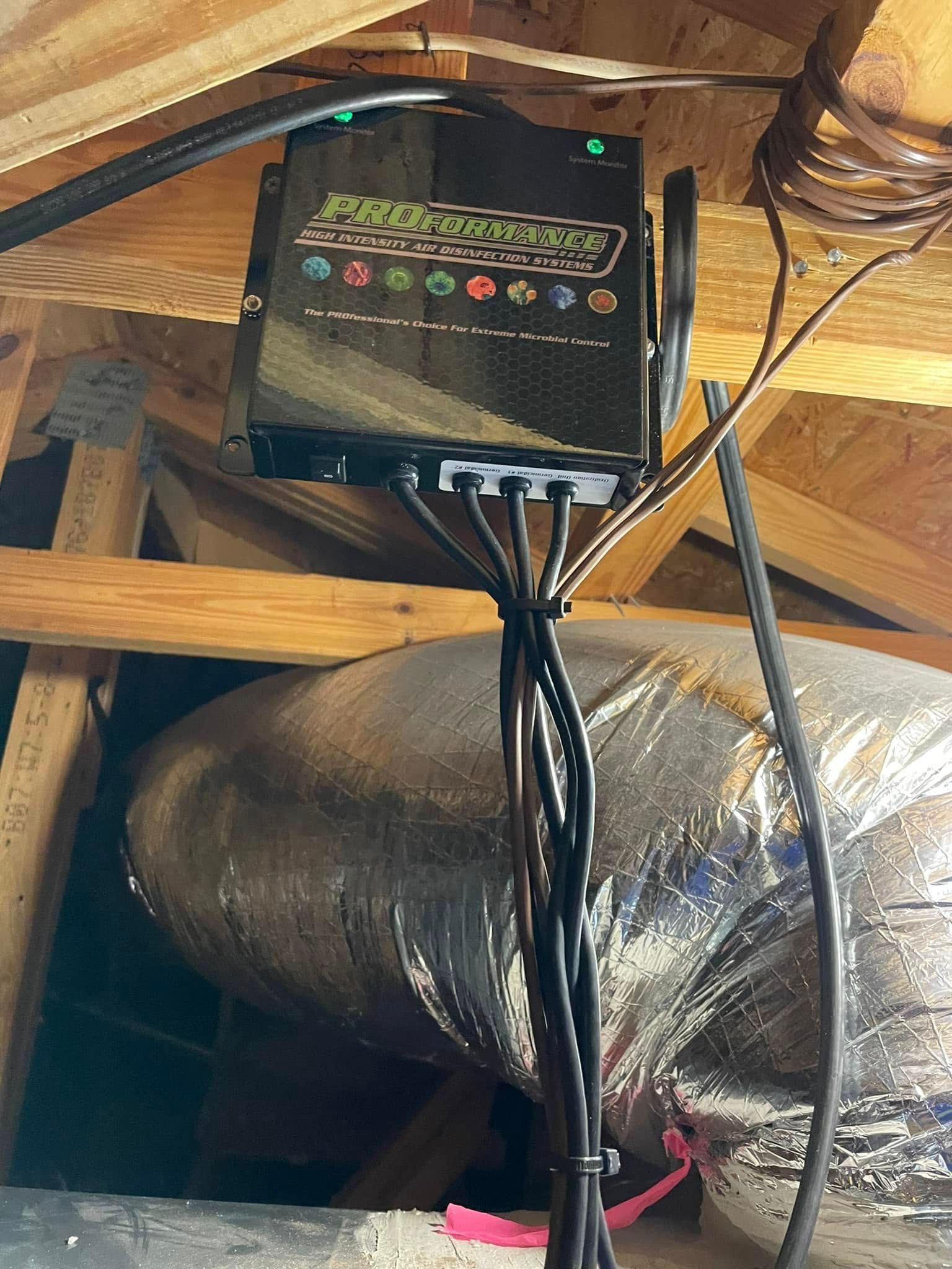 Black HVAC control box mounted on attic rafters above insulated ductwork, with cables hanging below