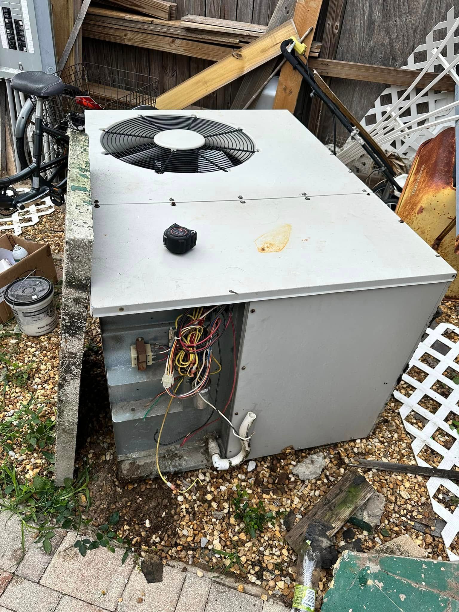 Gray outdoor HVAC unit with exposed wiring, sitting in a cluttered yard with debris and fencing.