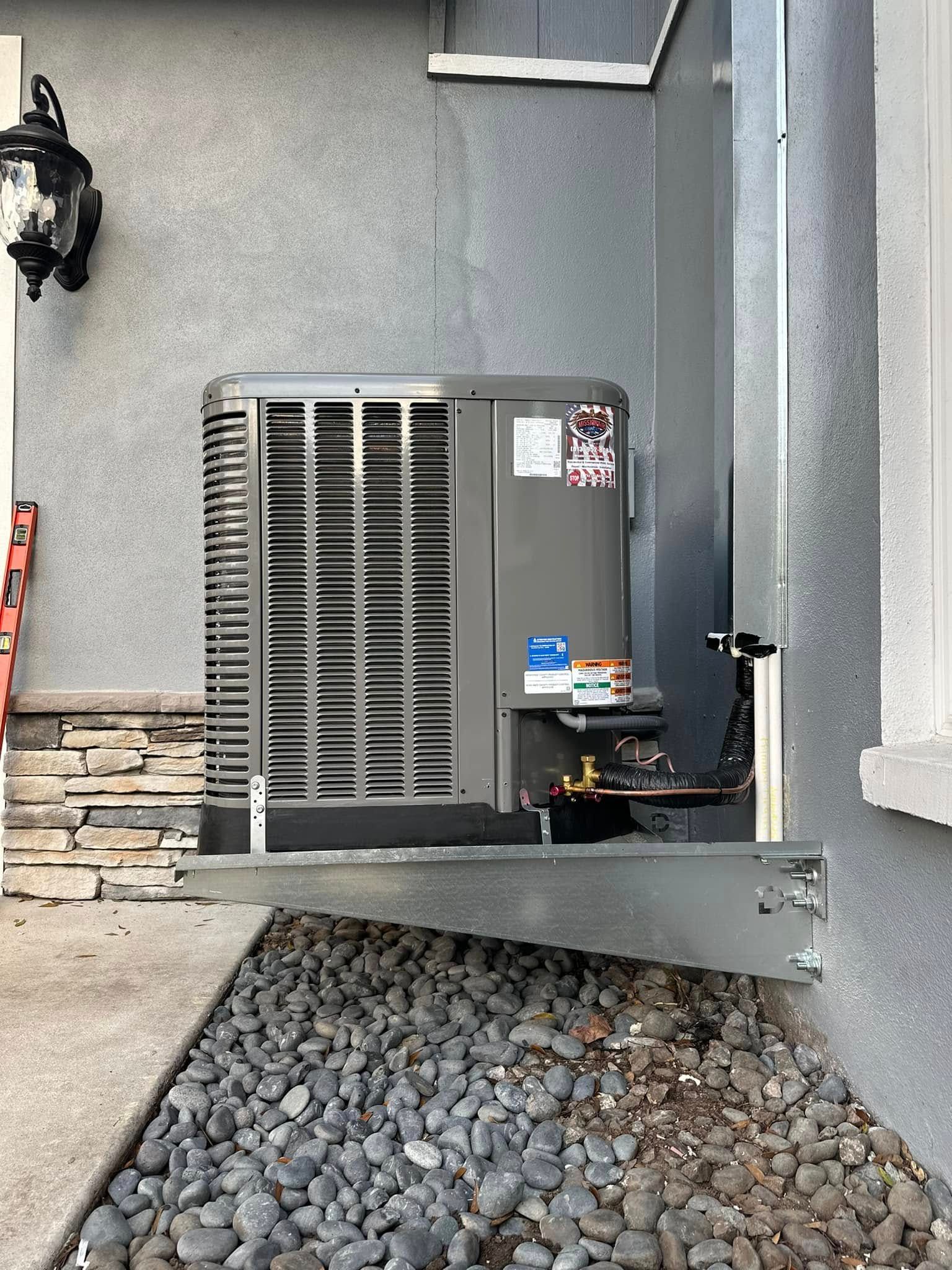Outdoor air conditioning unit beside a gray stucco wall, with gravel and stone landscaping below.