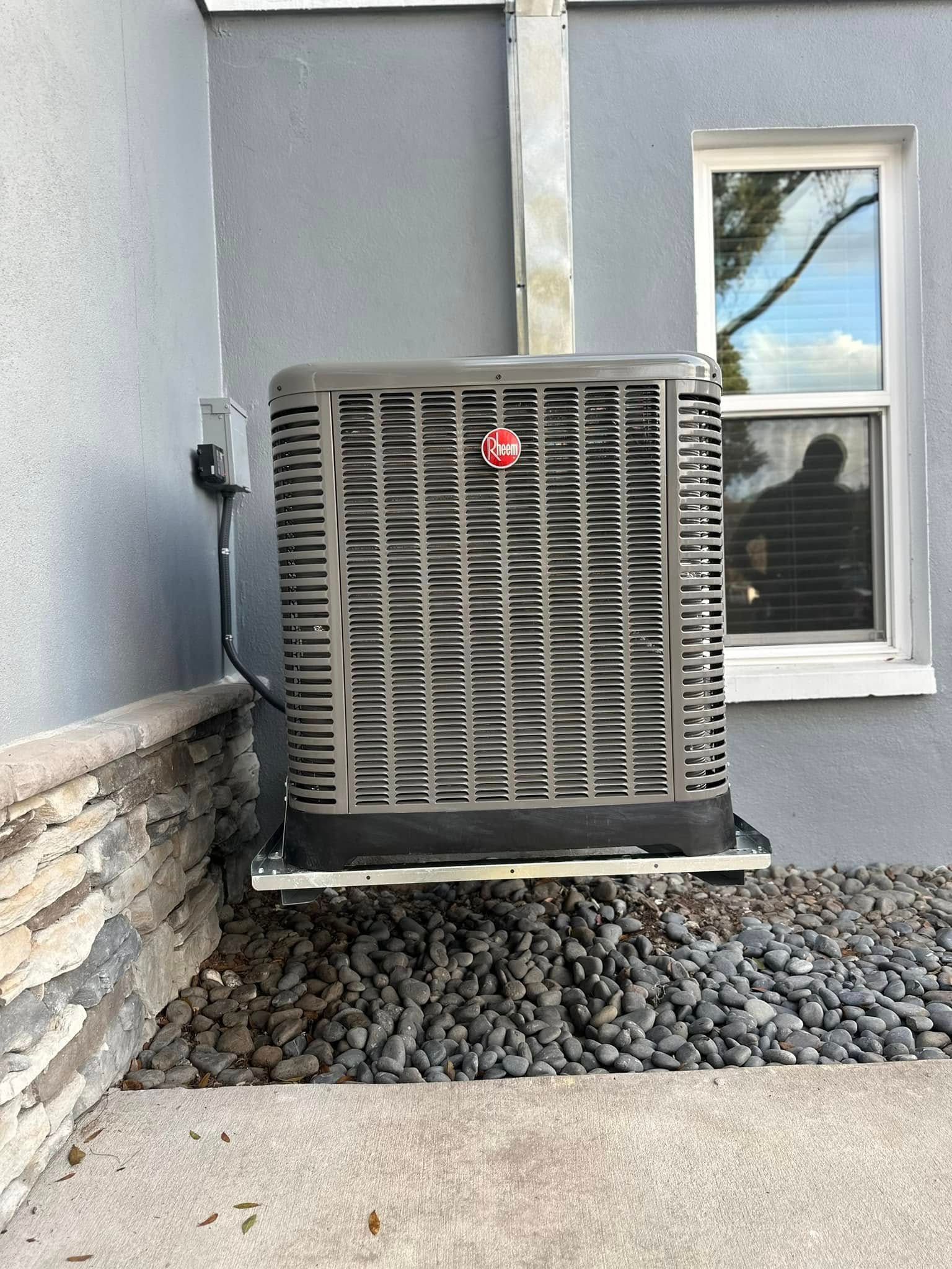 Outdoor HVAC unit beside a gray house wall and window, set on gravel near a stone foundation