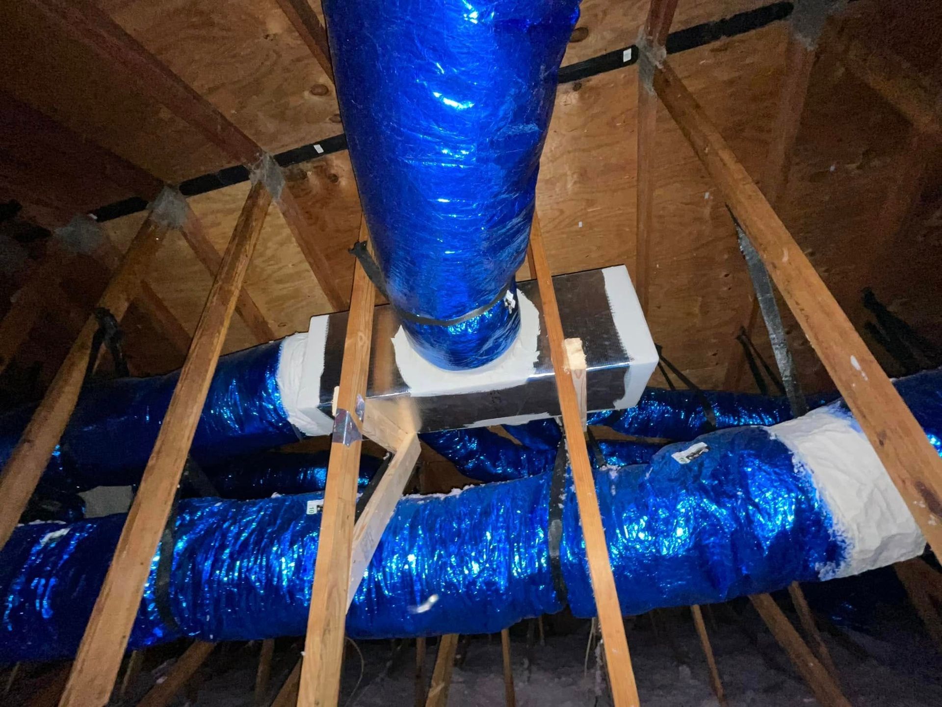 Blue insulated ductwork in an attic with wooden rafters and white support straps.