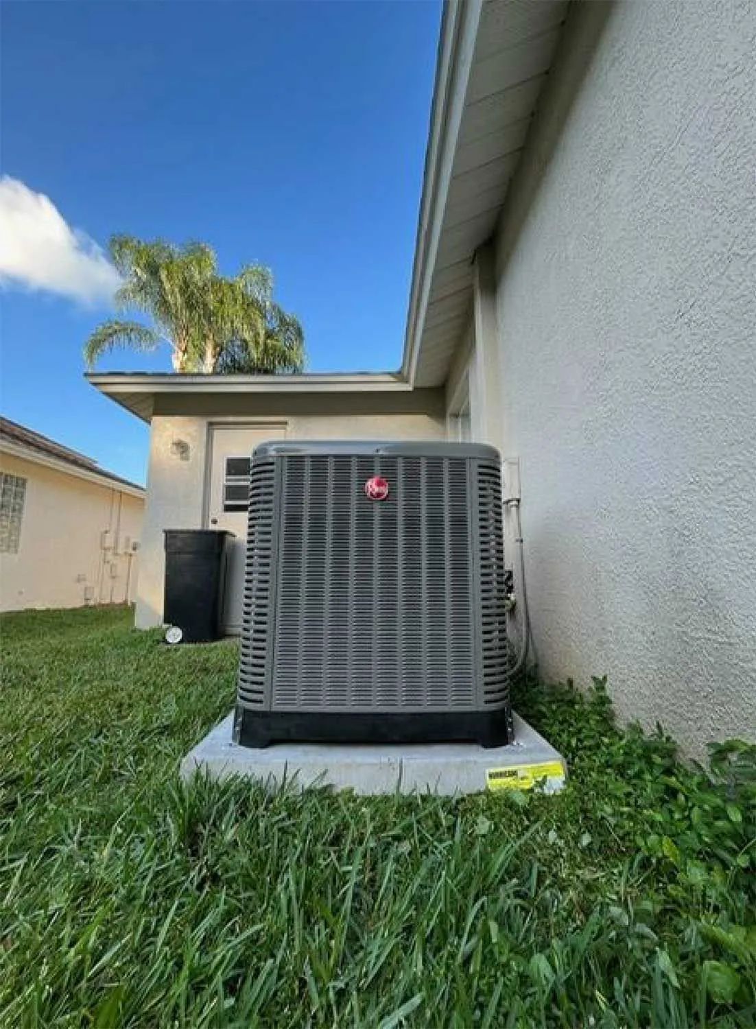 Outdoor air conditioner unit beside a house wall, on a concrete pad in a grassy yard under a blue sky