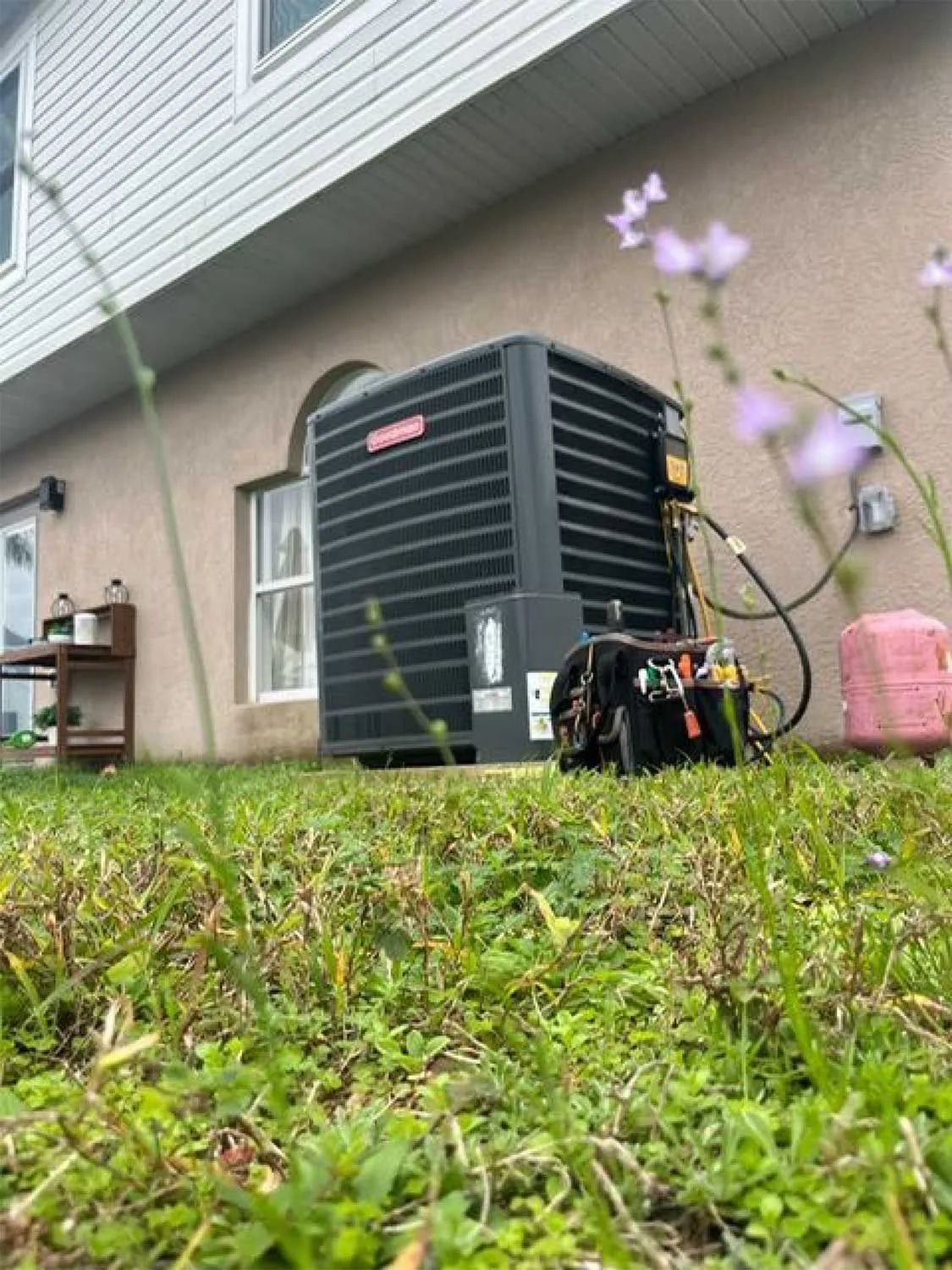 Outdoor HVAC unit beside a house, seen from low grass with a pink flower in the foreground.