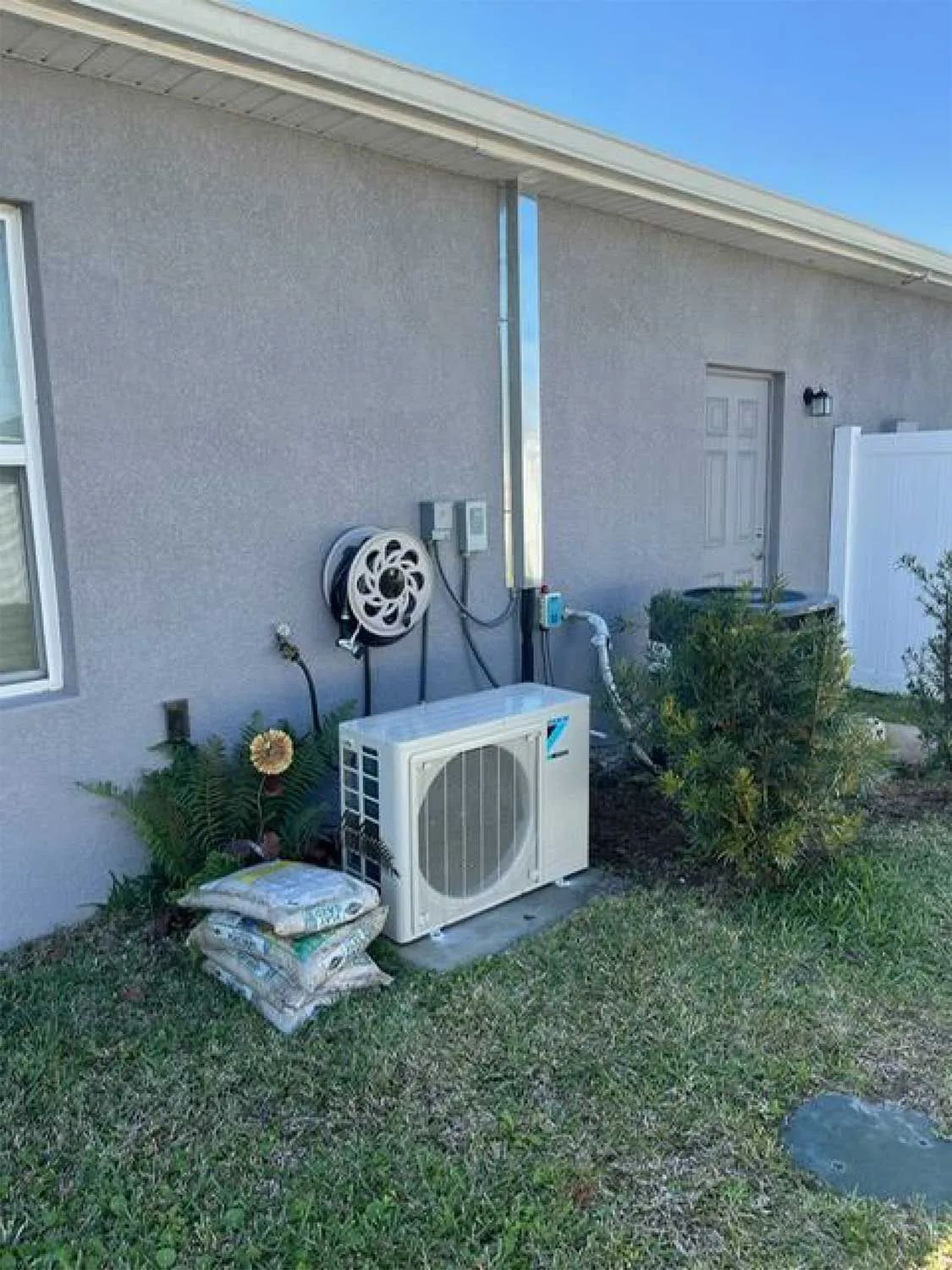 Outdoor HVAC unit beside a house wall with pipes, hose reel, and shrubs in a grassy yard
