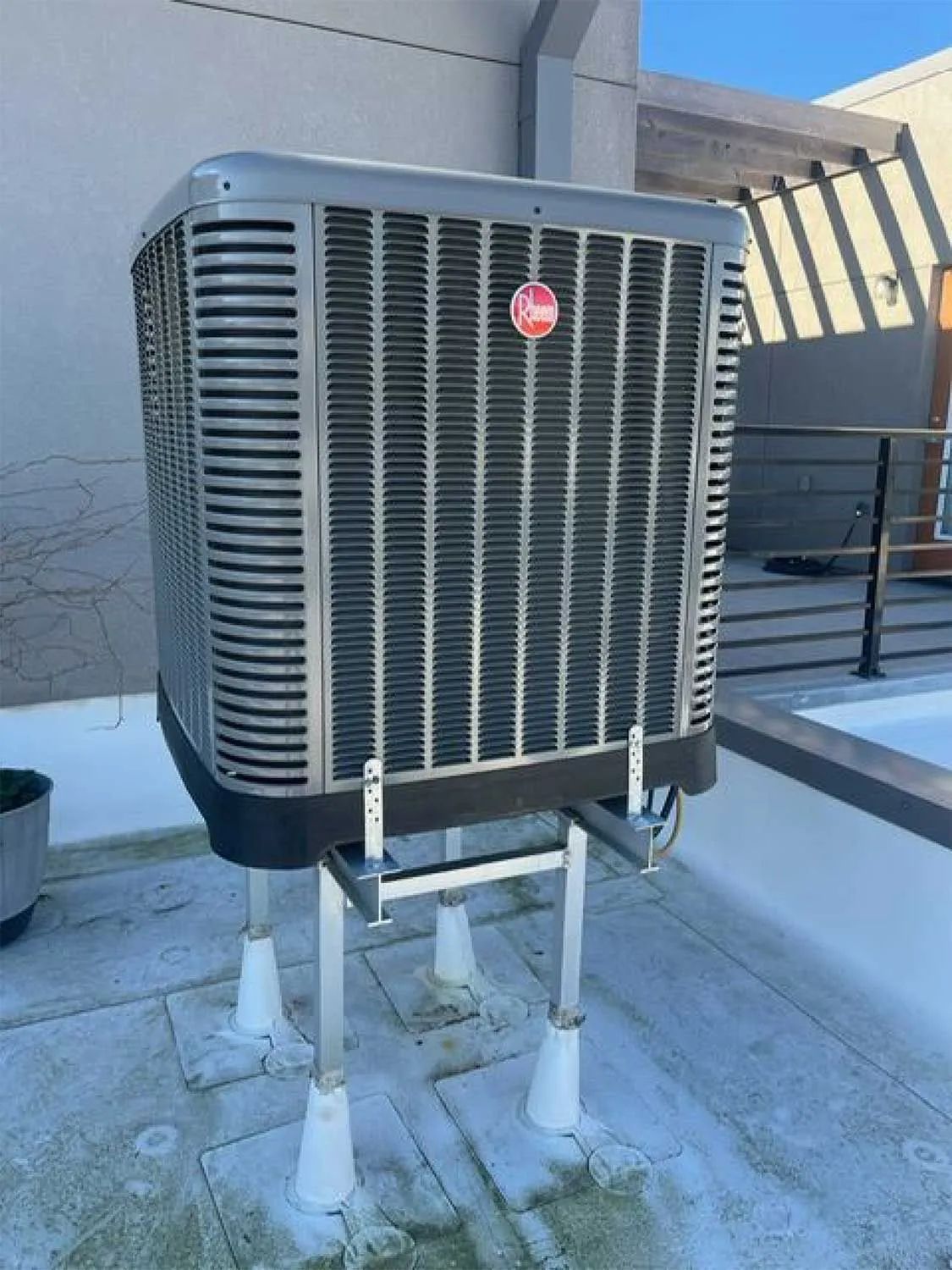 Outdoor rooftop HVAC unit with gray vents and red logo on white supports next to a building wall