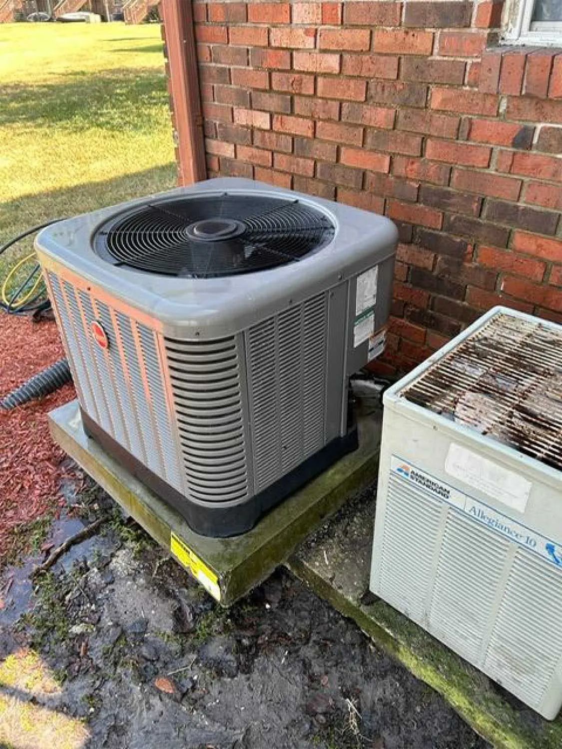 Outdoor air conditioner unit beside a brick wall, with a smaller unit nearby on a wet concrete pad.