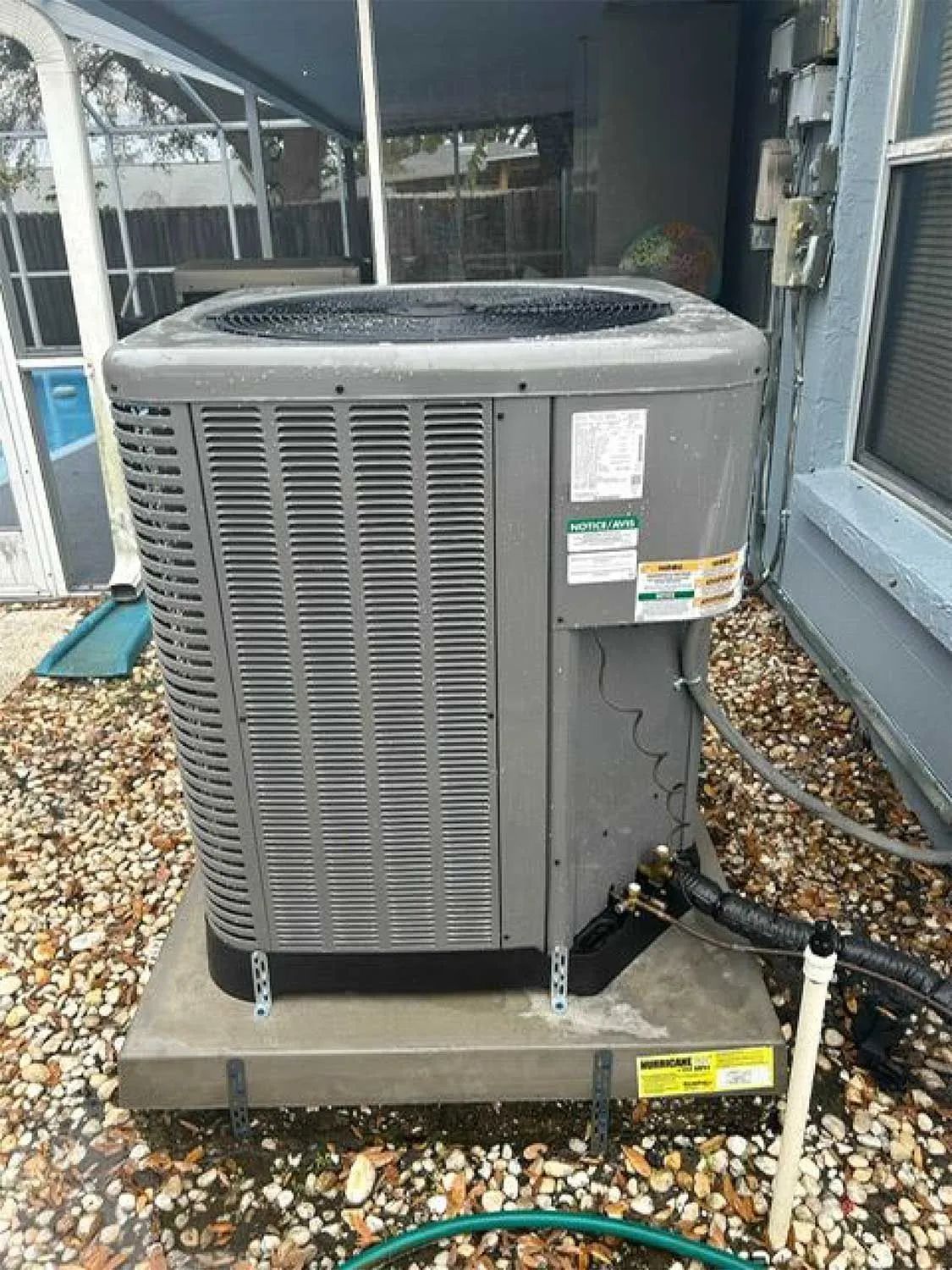 Outdoor central air conditioner unit on a concrete pad beside a house, with hoses and gravel nearby