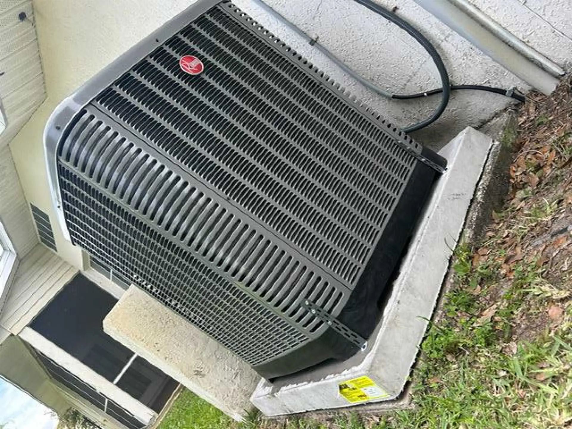 Outdoor air conditioner unit beside a house, with a red sticker on top and black hoses nearby