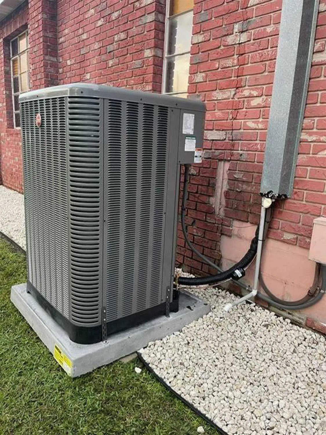 Outdoor air conditioner unit beside a brick house wall on a concrete pad, with gravel nearby