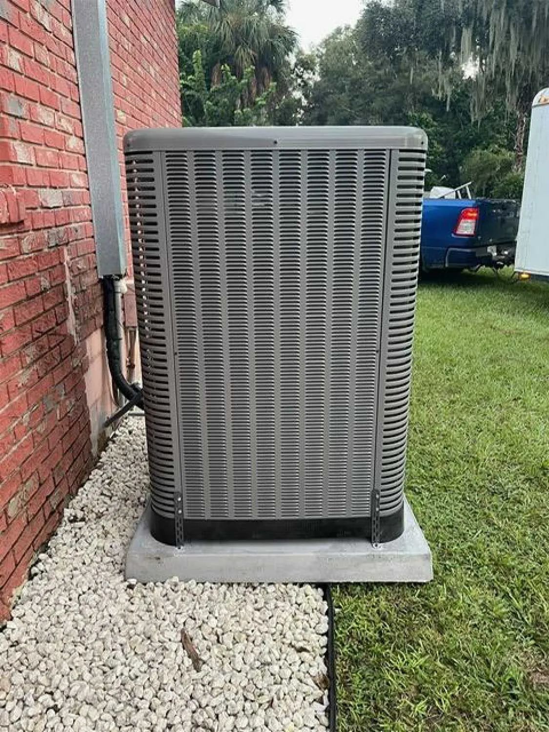 Outdoor gray HVAC unit beside a red brick wall, set on a concrete pad next to gravel and grass.