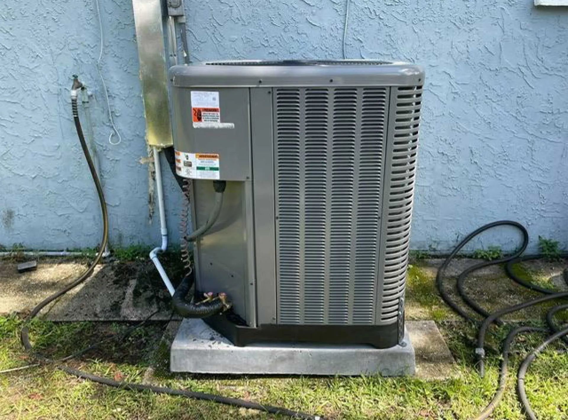 Outdoor air conditioner unit beside a blue house wall, with hose and concrete pad.