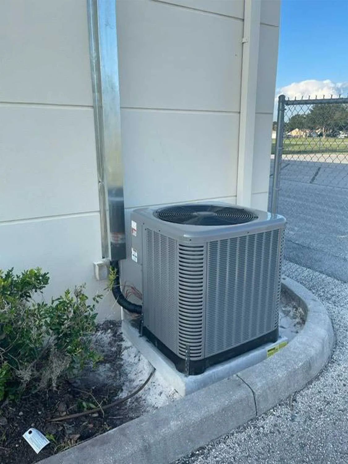 Outdoor HVAC unit beside a building wall, set in a curb-edged bed near a parking lot