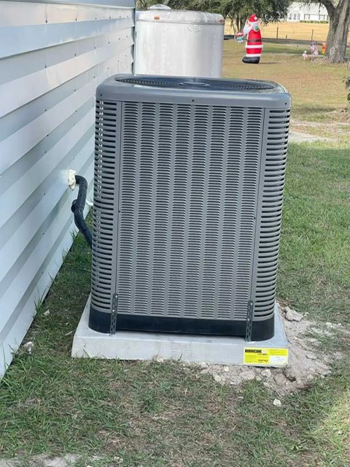 Outdoor HVAC unit beside a white house wall on a grassy yard, with a utility tank and traffic cone in the background