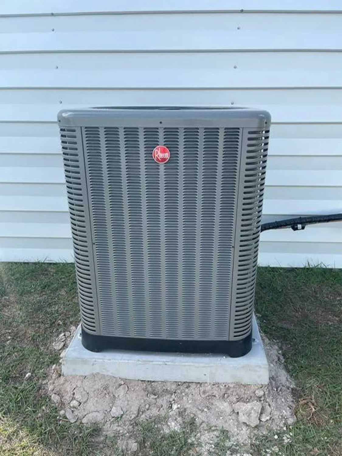 Outdoor air conditioner unit on a concrete pad against white siding with a black pipe on the right.