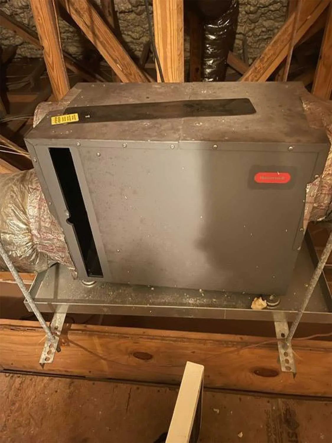 Gray HVAC furnace unit in an attic, sitting on a metal stand beneath exposed roof rafters.