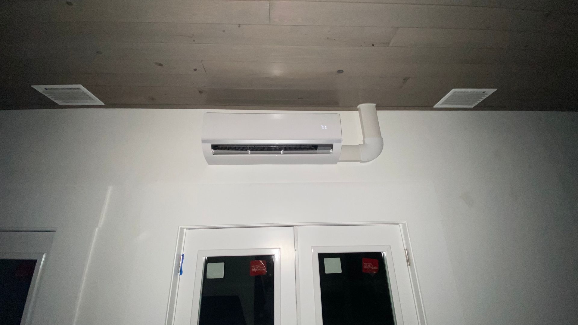 Wall-mounted air conditioner above two black-framed windows on a white wall, with ceiling vents above.