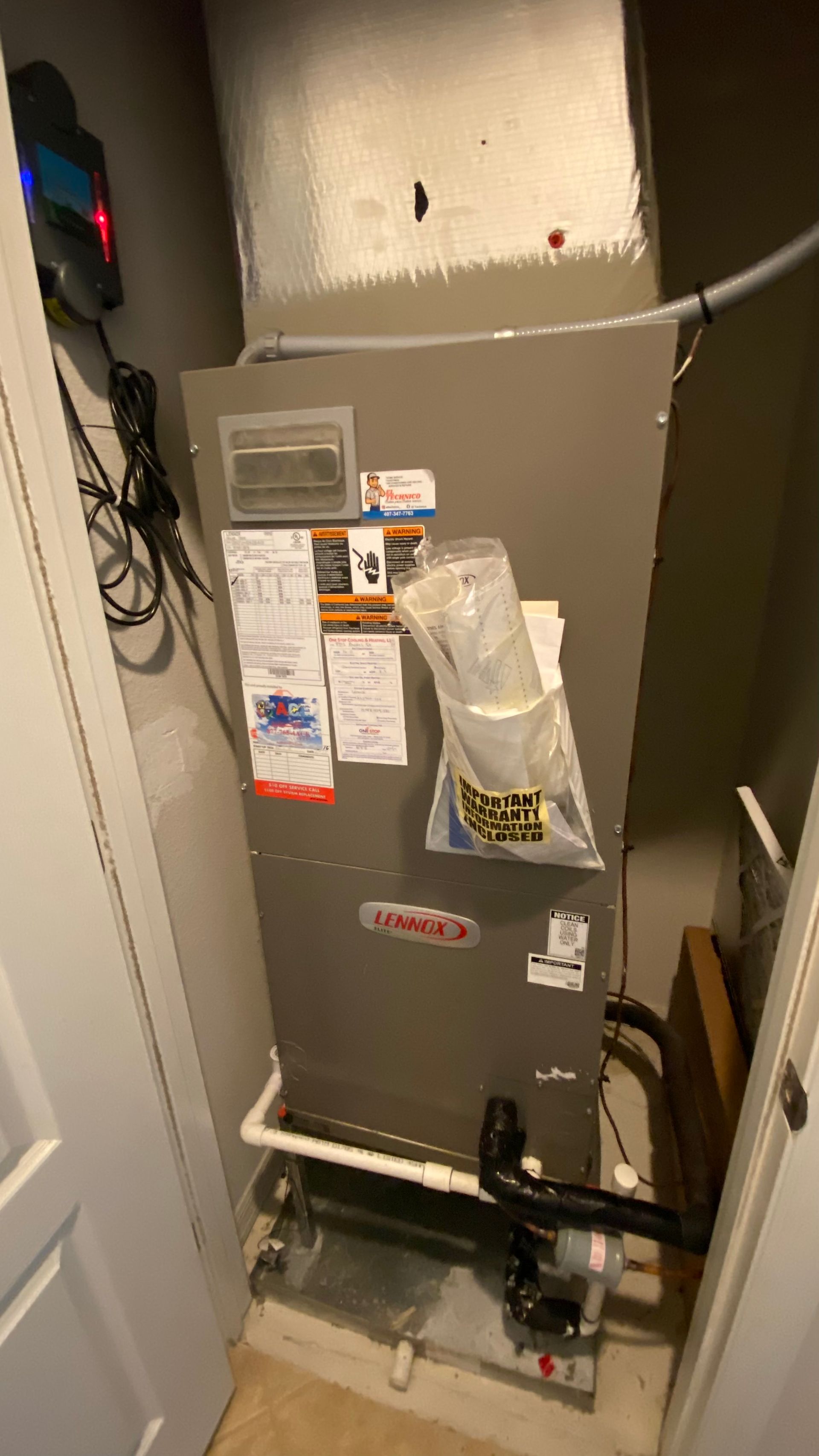 Gray HVAC furnace in a narrow closet with labels and pipes, beside a door and wall wiring.