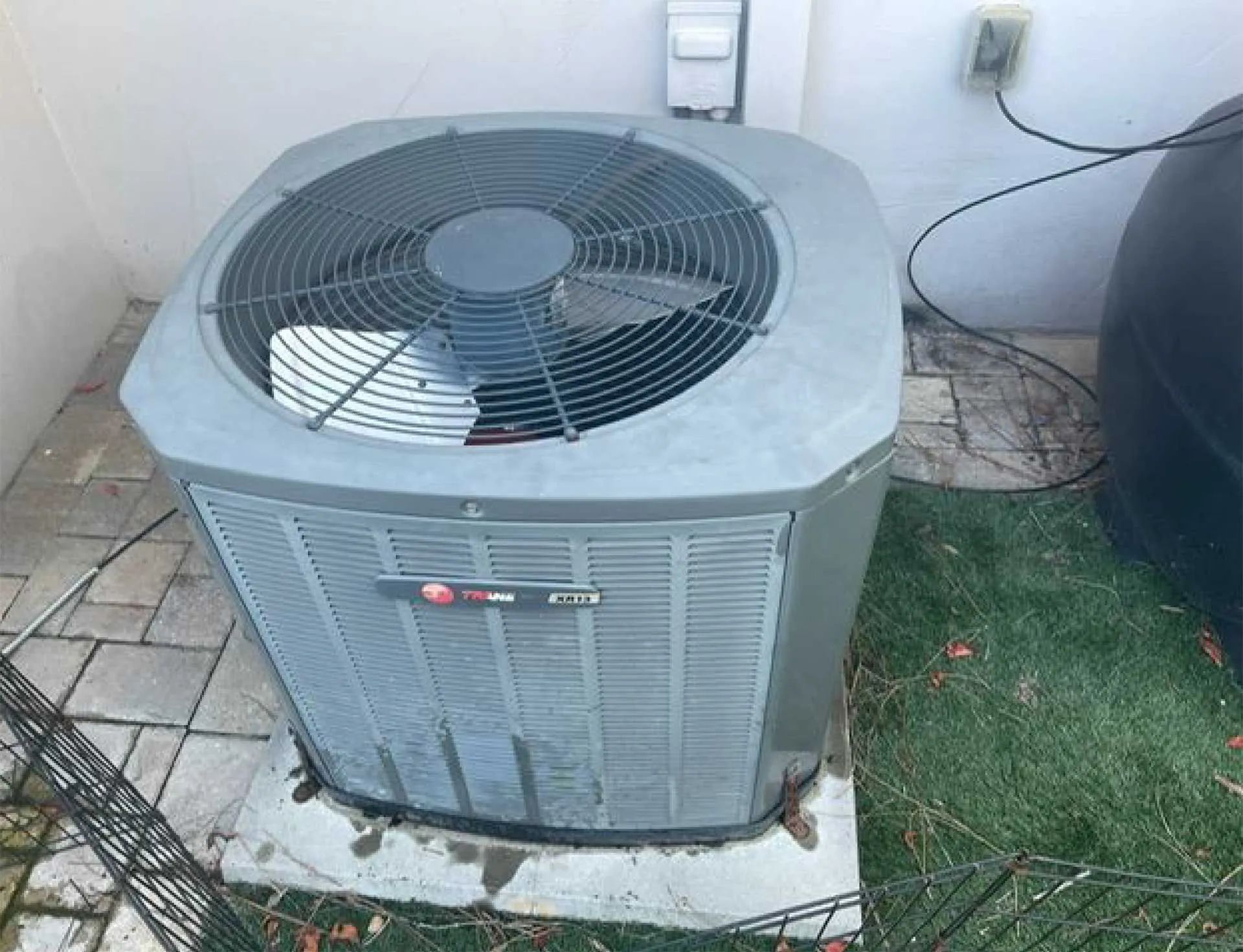 Gray outdoor air conditioner unit beside a wall, sitting on a concrete pad next to grass.