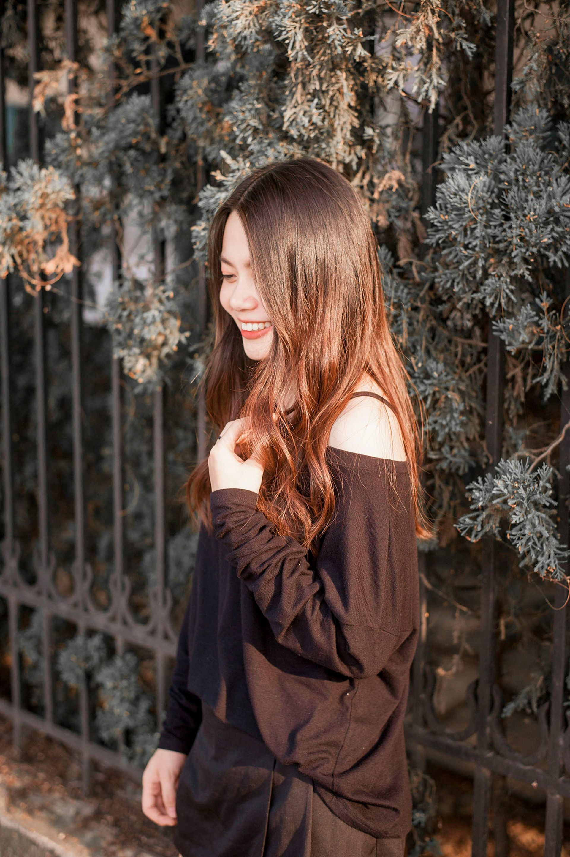 Young person in a black off-shoulder dress standing by an iron fence and greenery, smiling in warm light