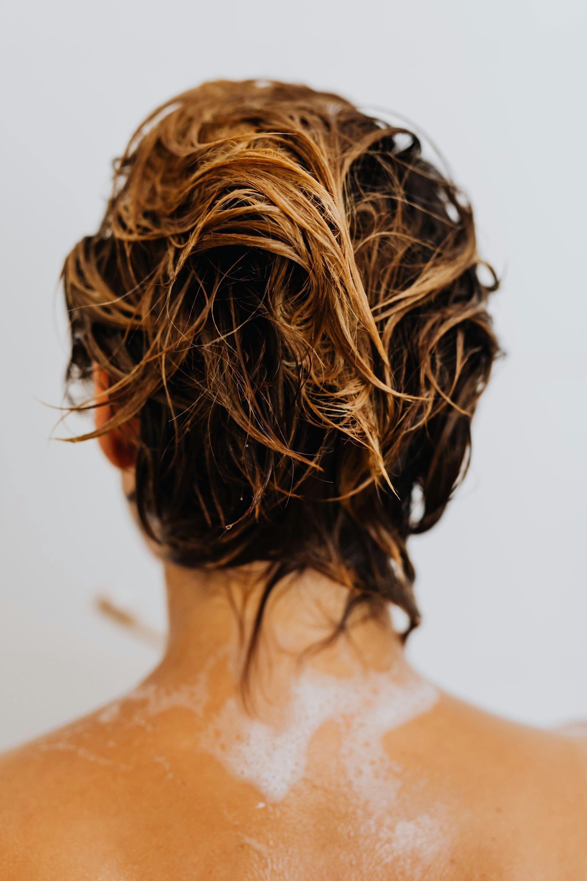 Back of a wet-haired person with sunlit strands, shoulders exposed, against a plain light background