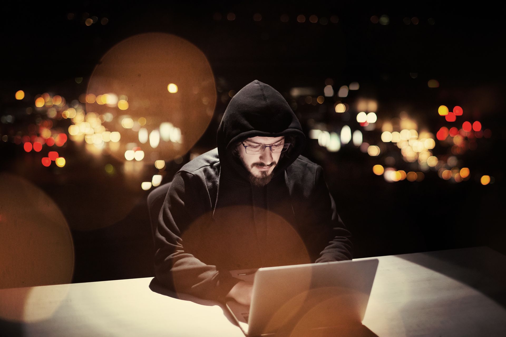 A man in a hoodie is sitting at a table using a laptop computer.