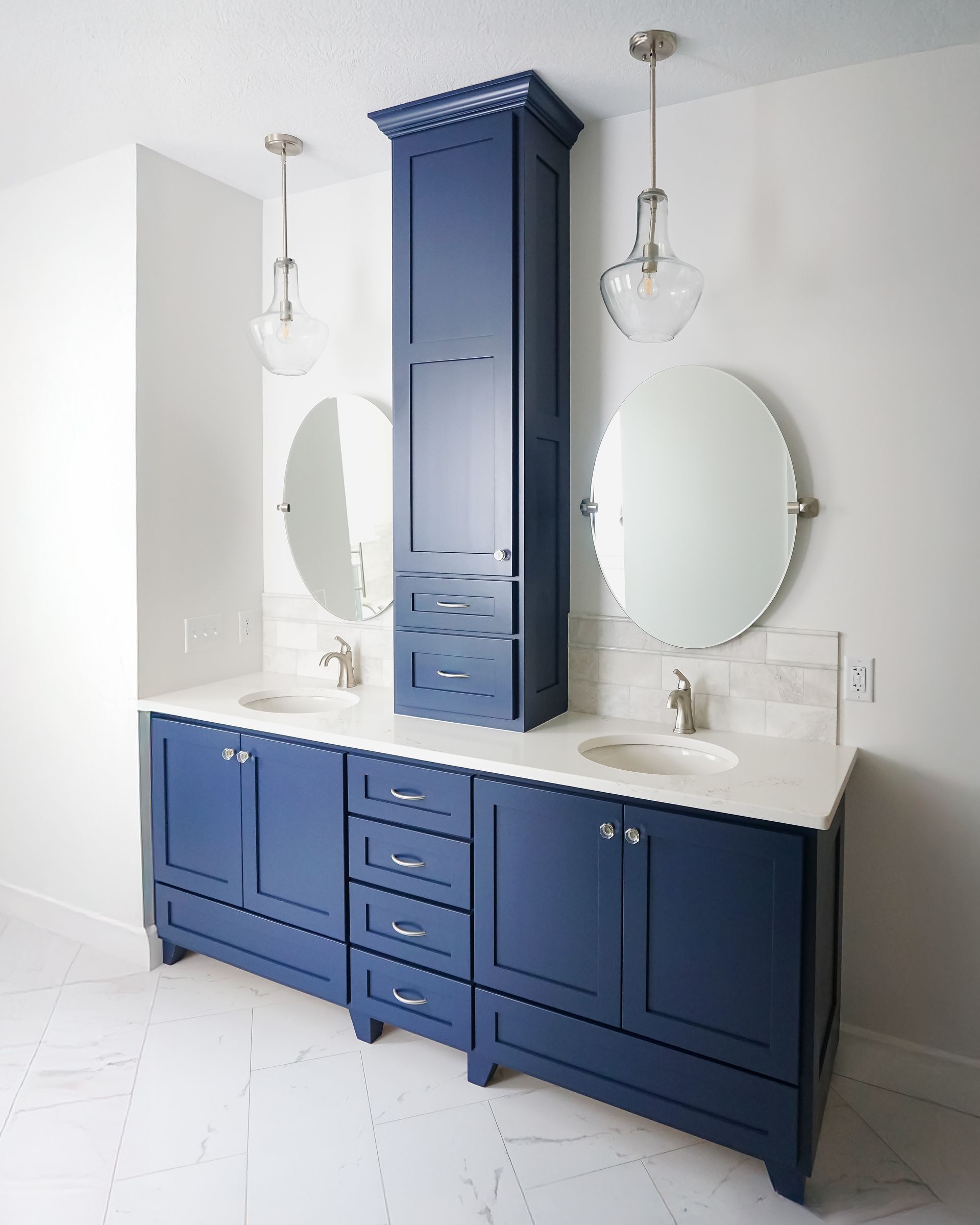 Blue double vanity with tall storage cabinet and oval mirrors in a white bathroom, lit by pendant lights.