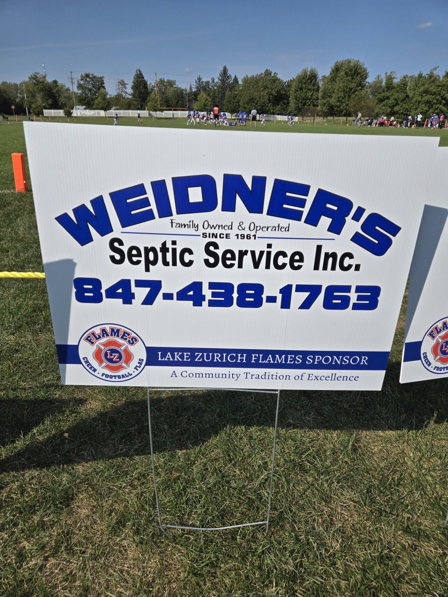 Sign for Weidner's Septic Service Inc., a Lake Zurich Flames sponsor, on a grassy field.
