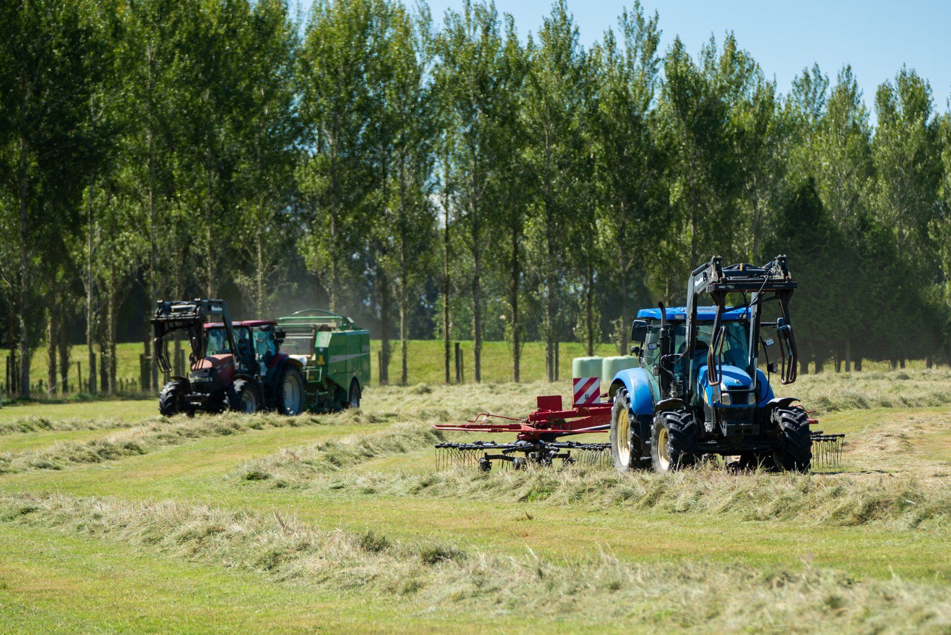 Farm machinery in foreground with a truck loaded with round hay bales in a grassy field.