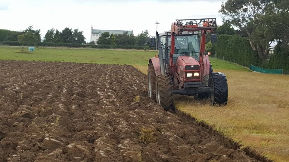 Red tractor plowing a muddy field beside a grassy area under cloudy skies