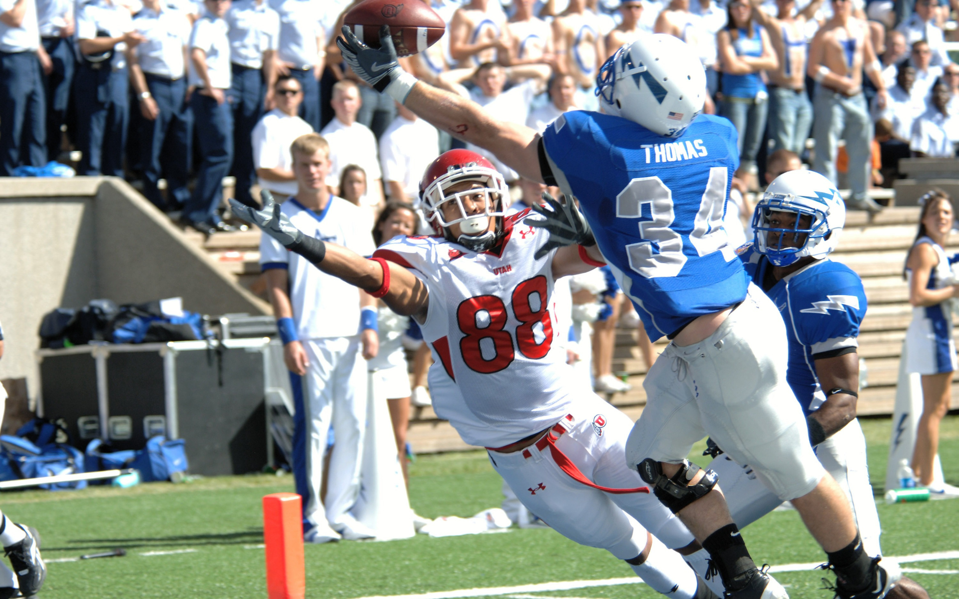 Football player in red leaping to catch a pass, blue player nearby; stadium background.