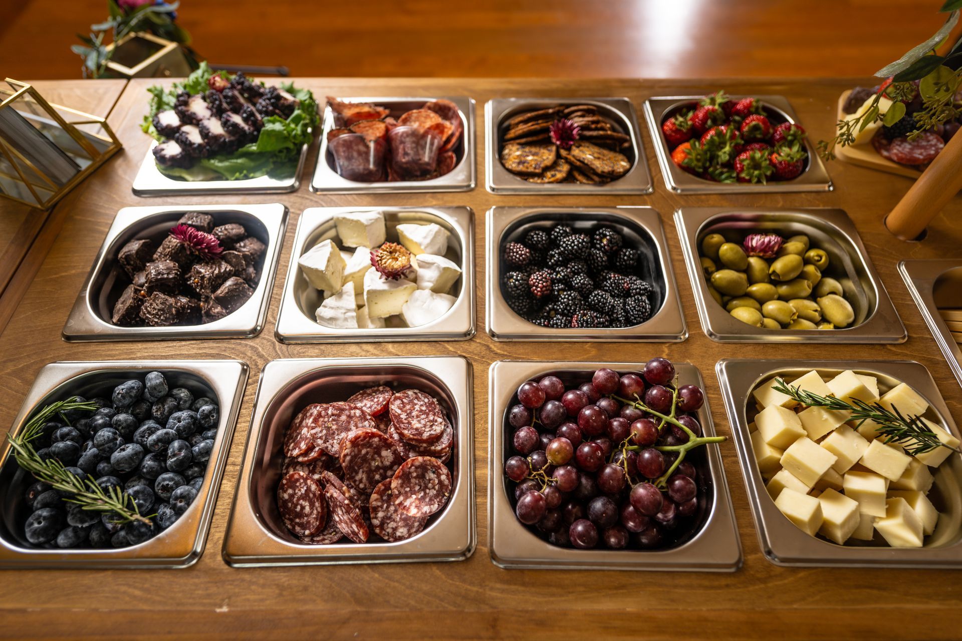 Assorted fruits, cheeses, and desserts arranged in metal trays on a wooden table.