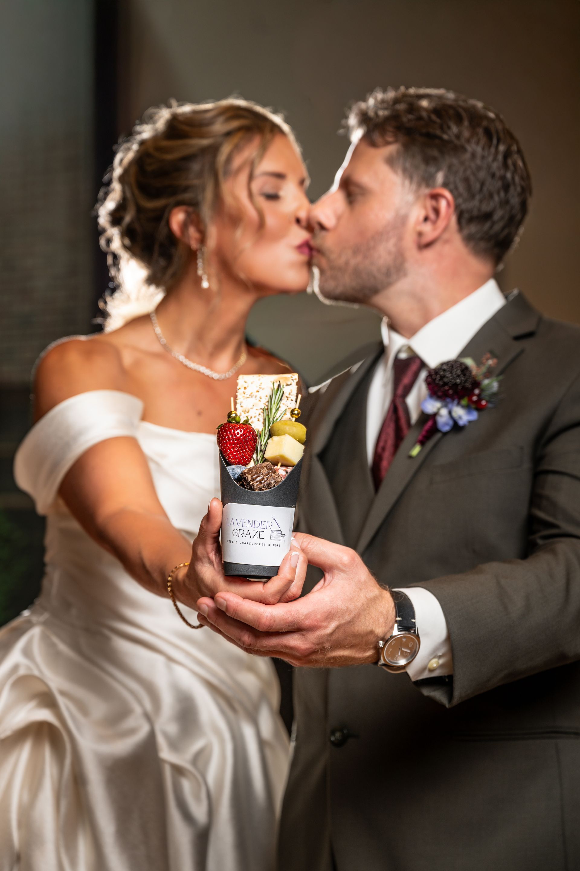 Wedding couple kissing while holding a decorated bottle, with a dark backdrop.