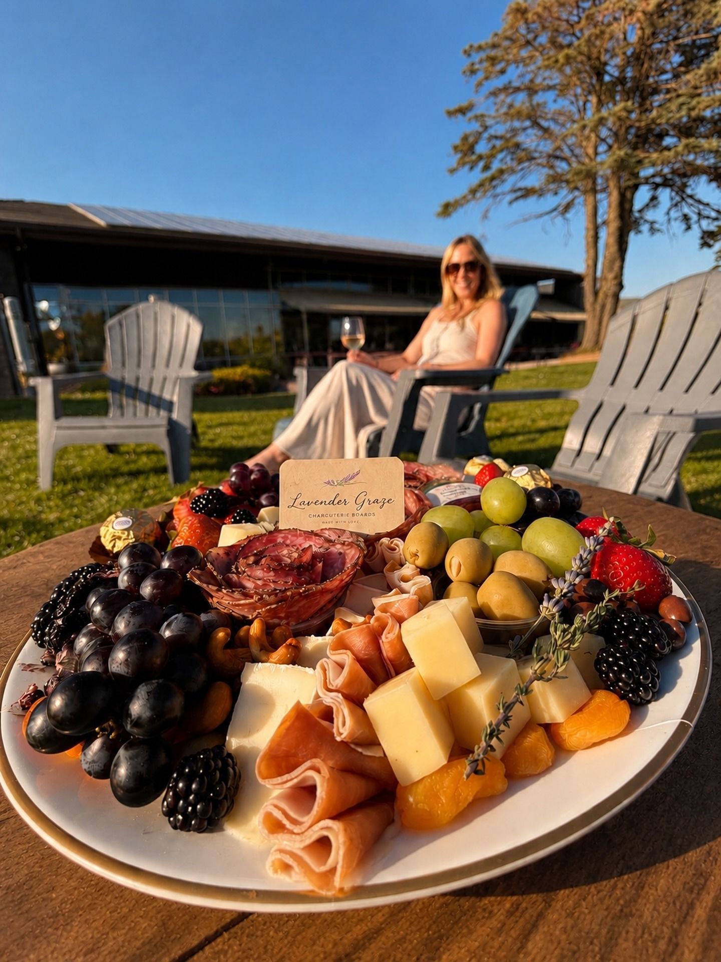 Fruit and cheese platter on a table outdoors, with a woman sitting in the background by the lake