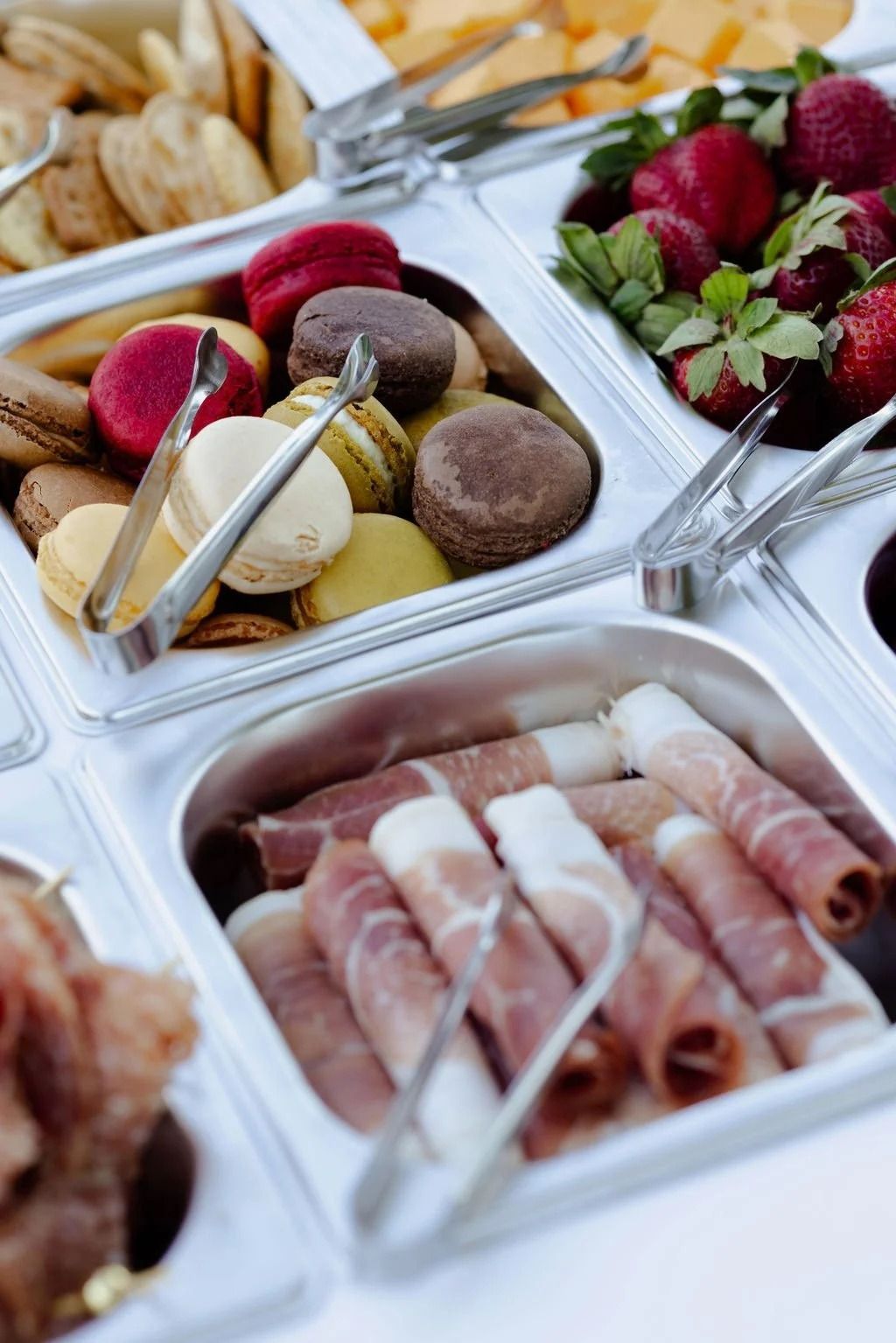 Buffet table with wrapped sandwiches, cheese, strawberries, chocolate desserts, and tongs